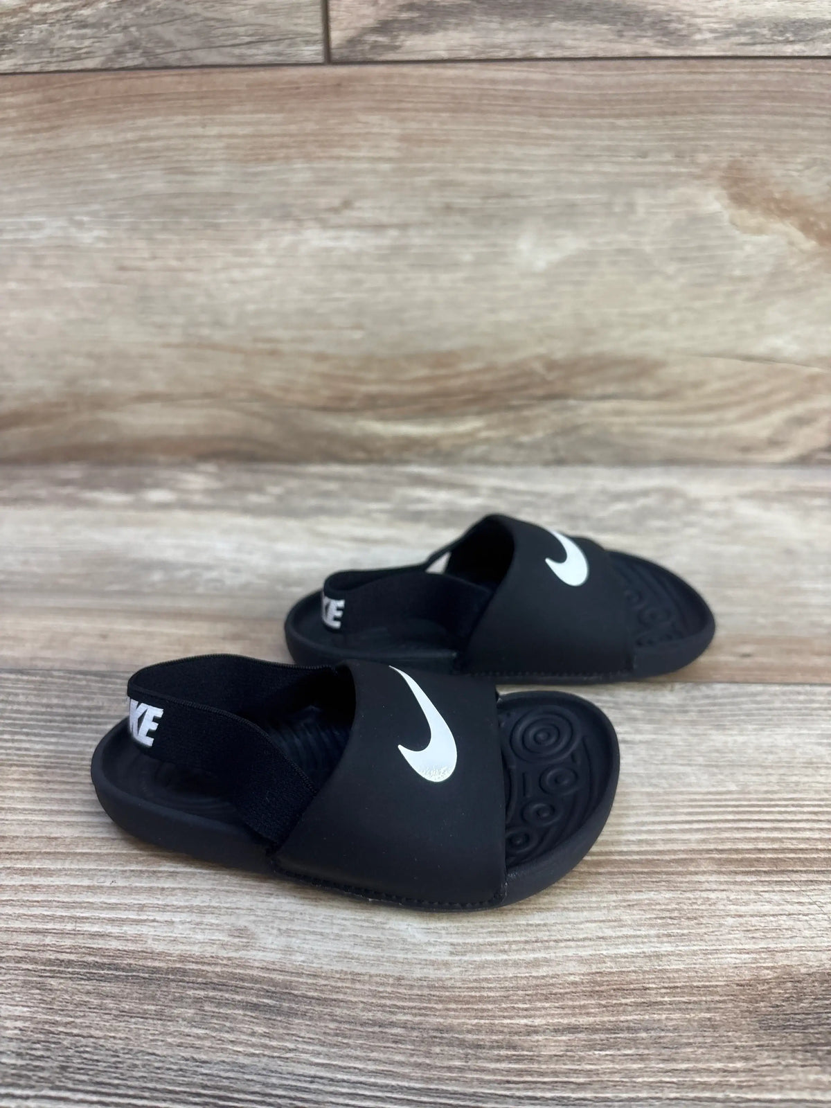 Angled view of Nike Kawa toddler black slide sandals showcasing white Swoosh and elastic heel strap on wood background