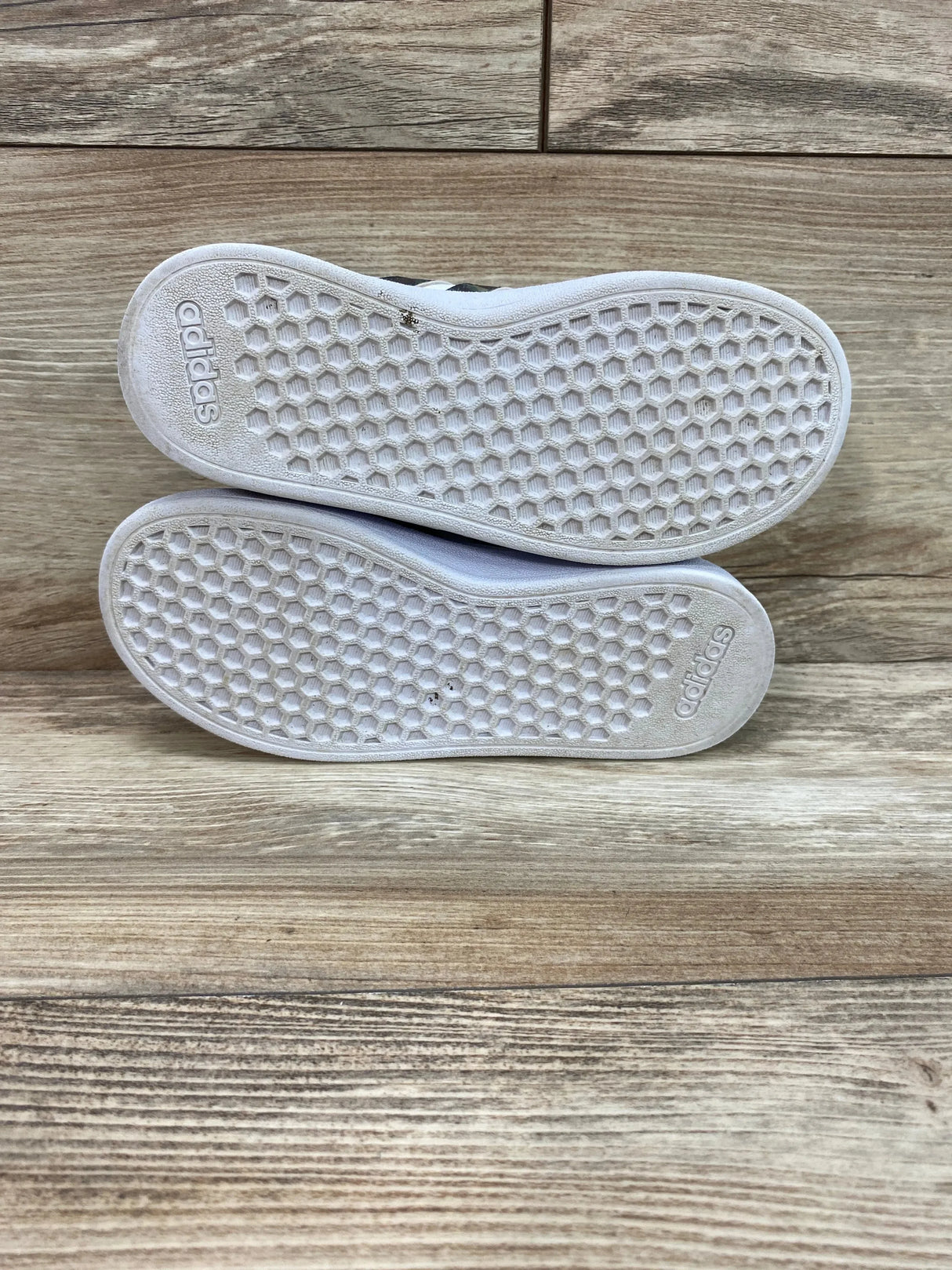 Bottom view of Adidas Neo Grand Court Base toddler shoes showing white honeycomb soles with adidas logo on wood background