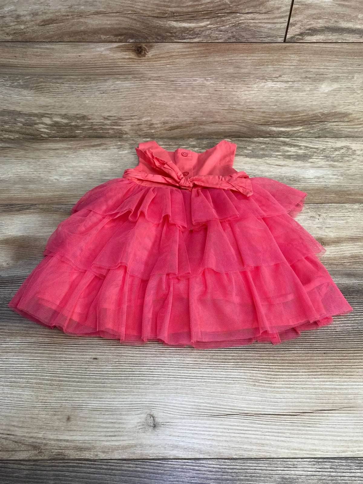 Flat lay of Heirlooms tulle baby dress in pink showing tiered ruffle skirt and waist bow on wood background