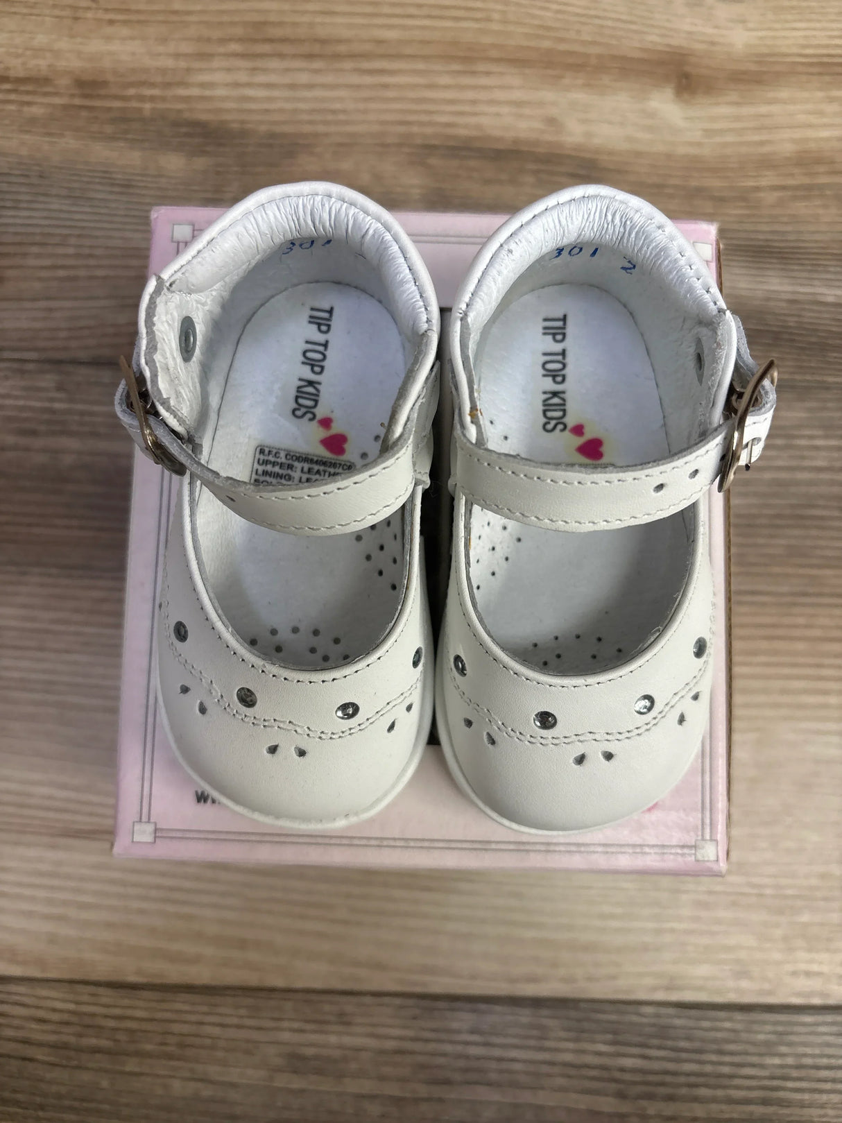 Pair of white leather baby shoes with buckle straps and decorative cutouts on a wooden surface