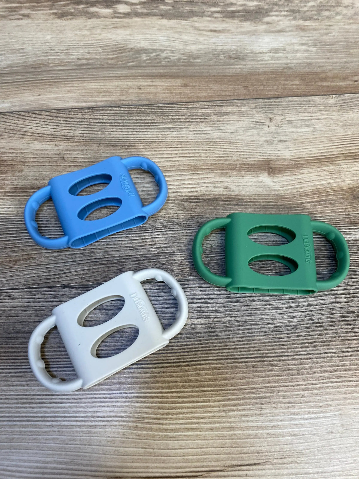 Top-down view of three Dr Brown’s silicone narrow baby bottle handles in blue, green, and white showing double-loop grips and oval cutouts on wood surface