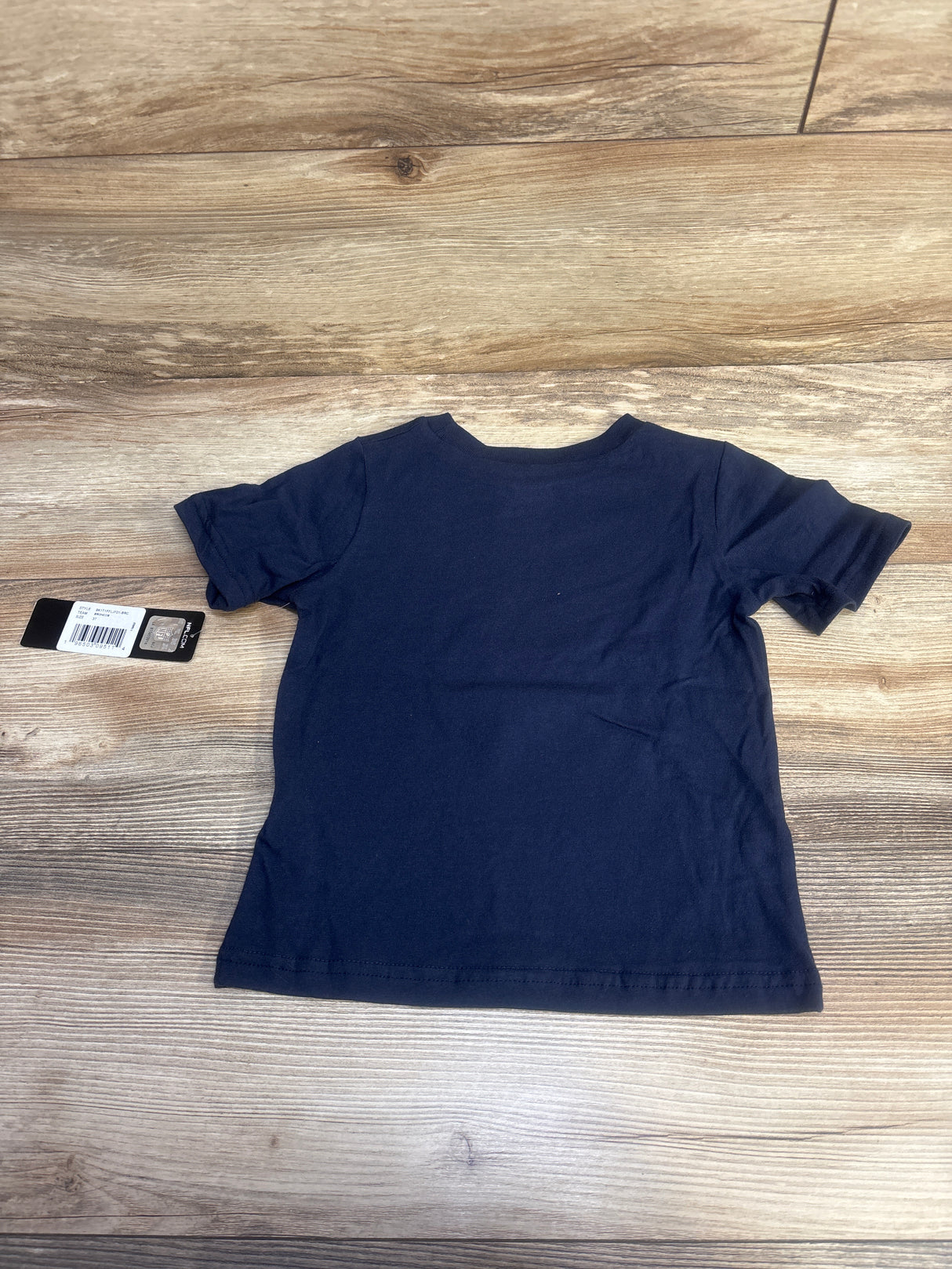 Back view of a plain navy blue short-sleeve toddler t-shirt on wooden floor with attached tag