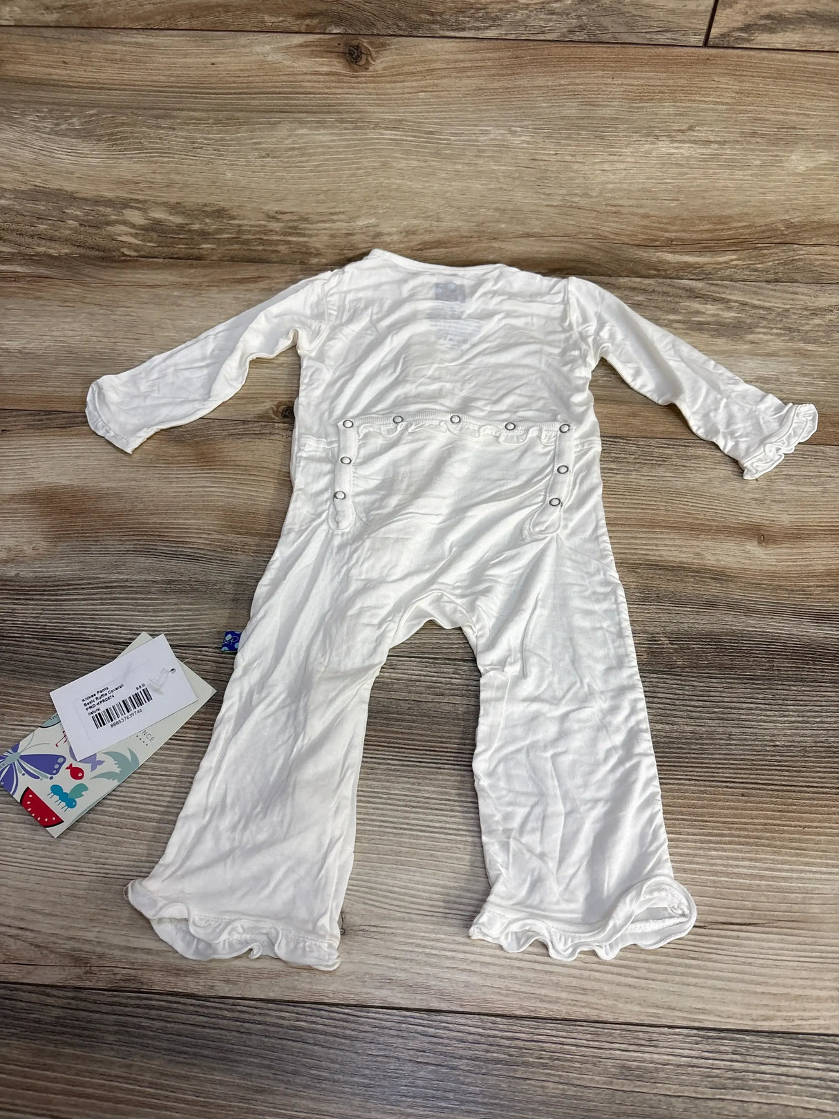 Back flat lay of white Kickee Pants ruffle coverall showing rear snap closure and ruffled cuffs on wood background