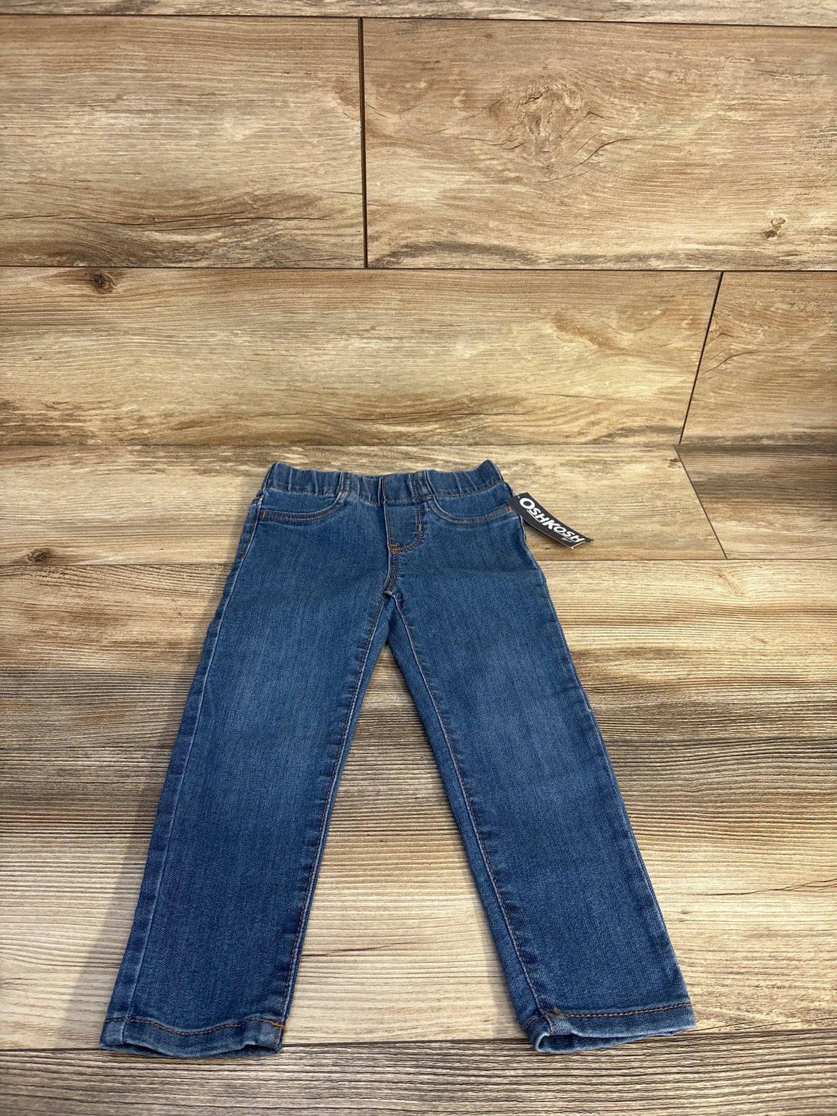 Front view of New OshKosh medium wash pull-on jeggings showcasing elastic waist and full-length legs on a wood background