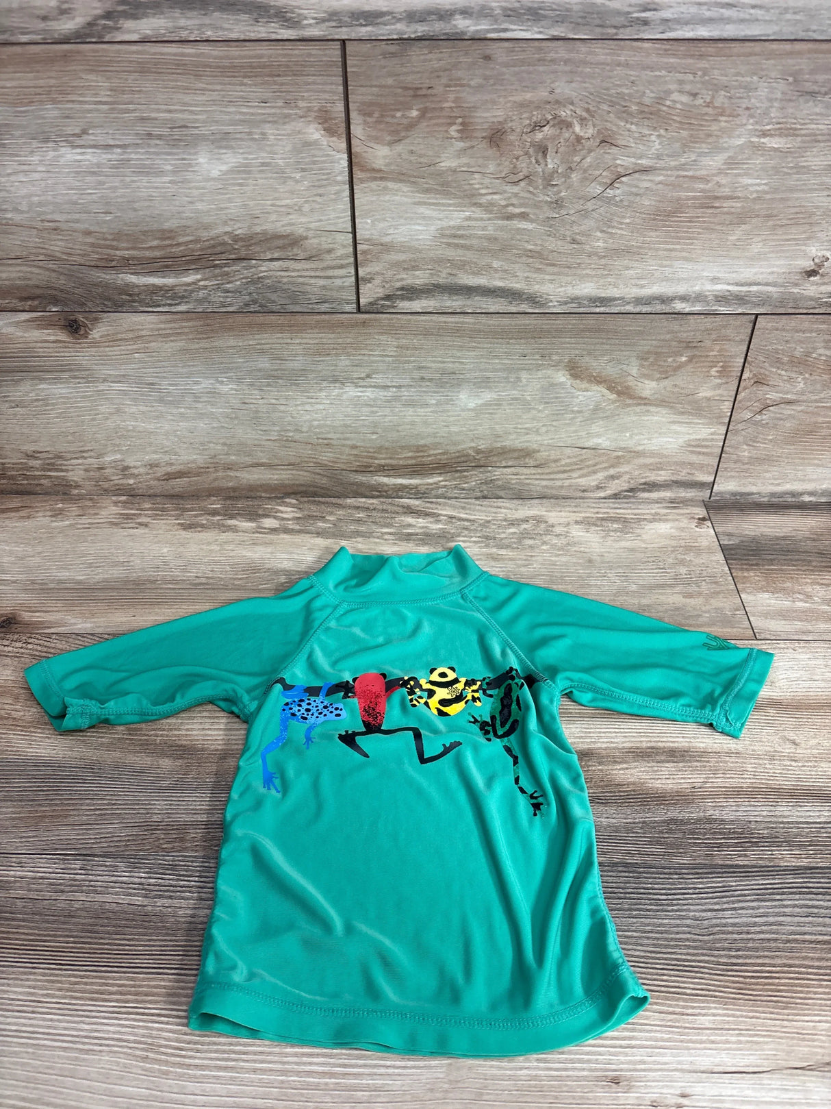 Front view of green UV Skinz Frog Rash Guard Shirt showcasing long sleeves, high collar, and frog graphic on a wood-look floor