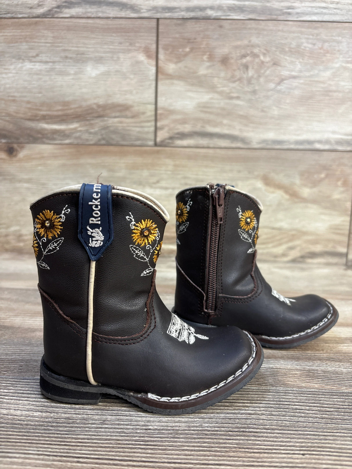 Brown toddler cowboy boots with sunflower embroidery, side zipper, and brand tag on wood floor