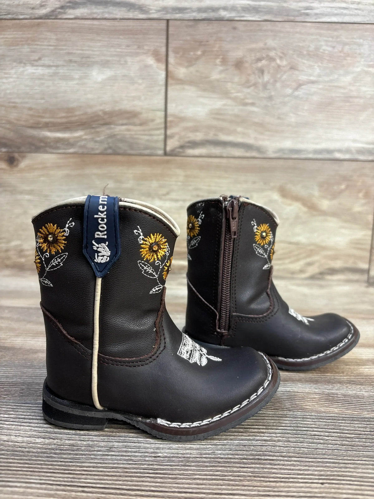 Brown toddler cowboy boots with sunflower embroidery, side zipper, and brand tag on wood floor