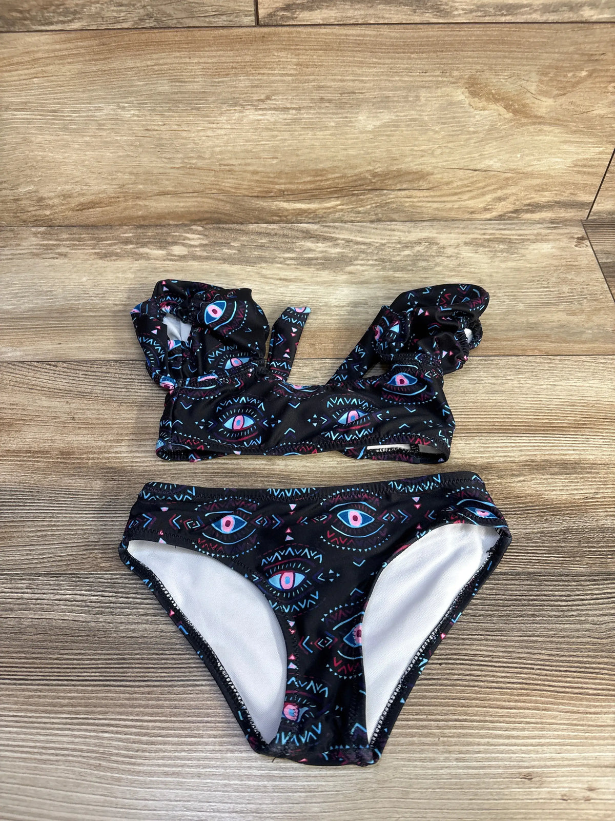 Front flat lay of 2T two-piece black evil eye swimsuit with ruffled shoulder straps and matching bottoms on wood floor