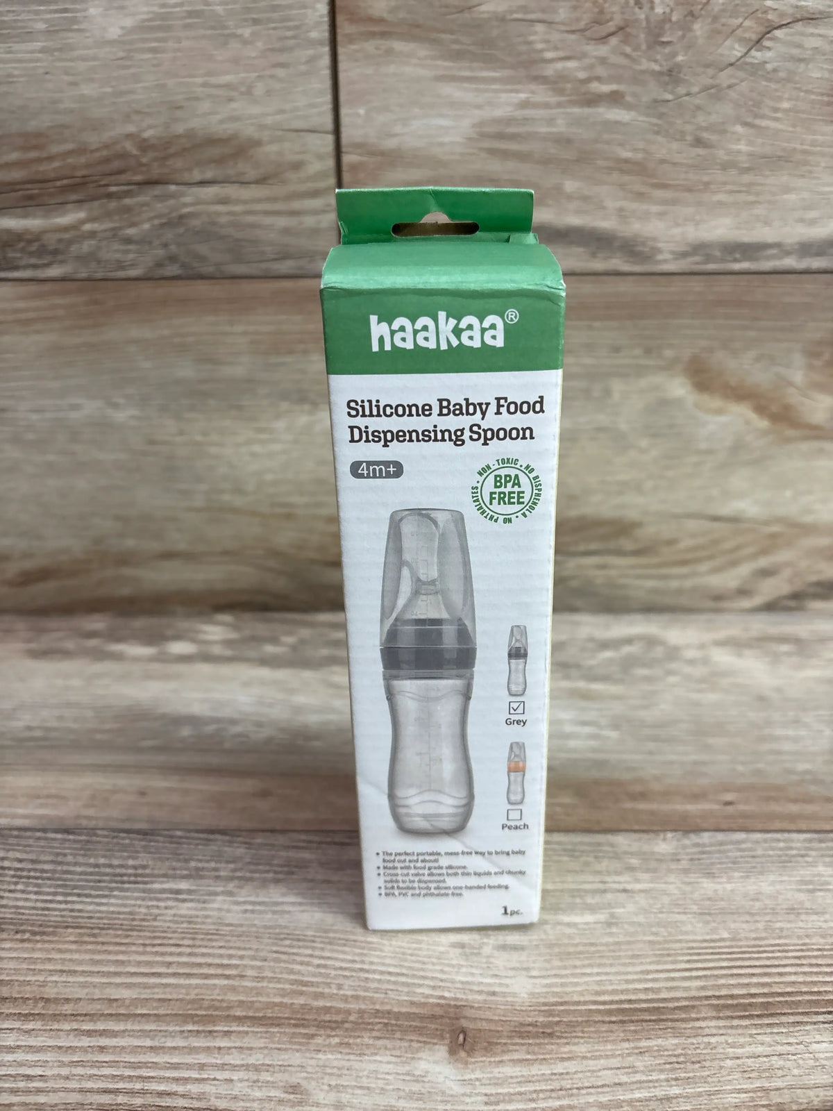 Front view of Haakaa silicone baby food dispensing spoon showcasing the gray color and 4m plus label in packaging on a wood surface