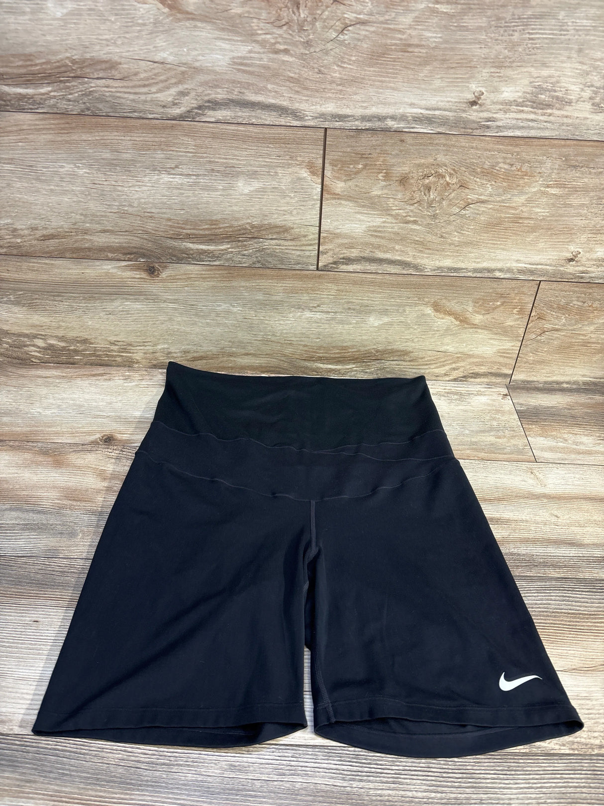 Flat lay of Nike maternity Dri-Fit black active shorts showing high fold-over maternity waistband and white Swoosh on wood floor background
