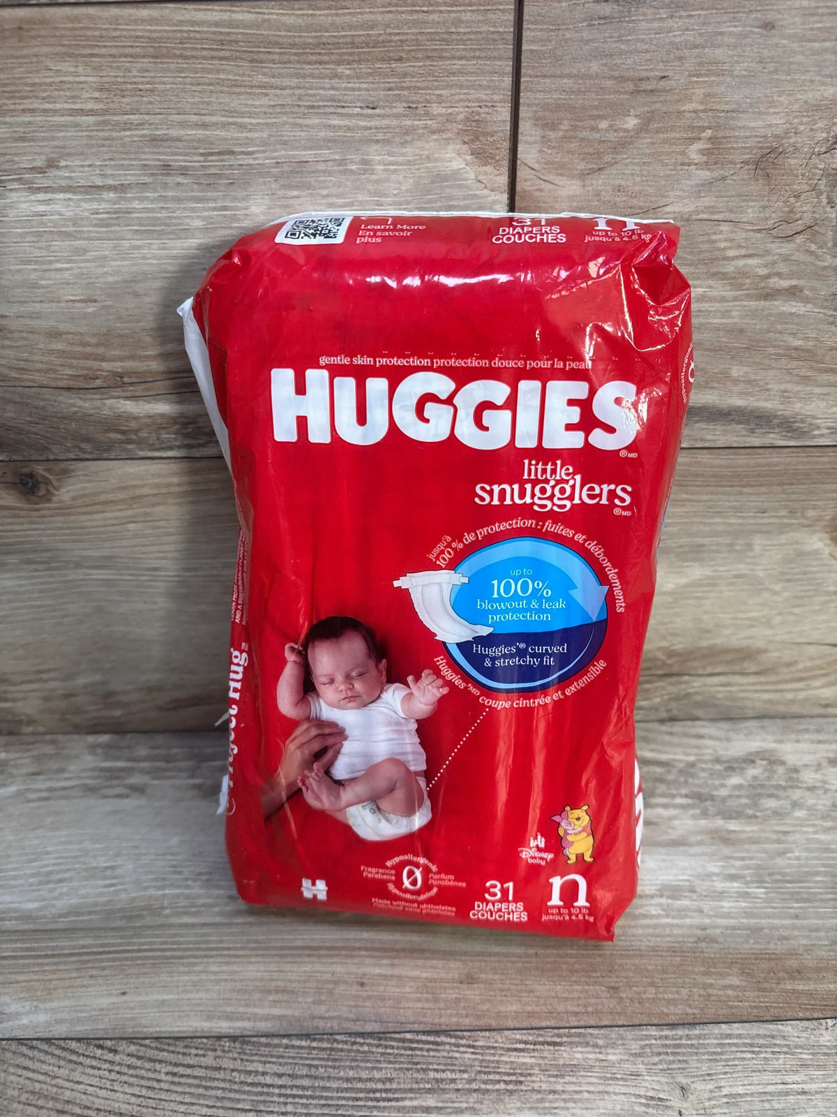 Front view of Huggies Little Snugglers newborn 31 count diaper pack showing red packaging, baby photo, and up to 100% blowout & leak protection badge on wood background