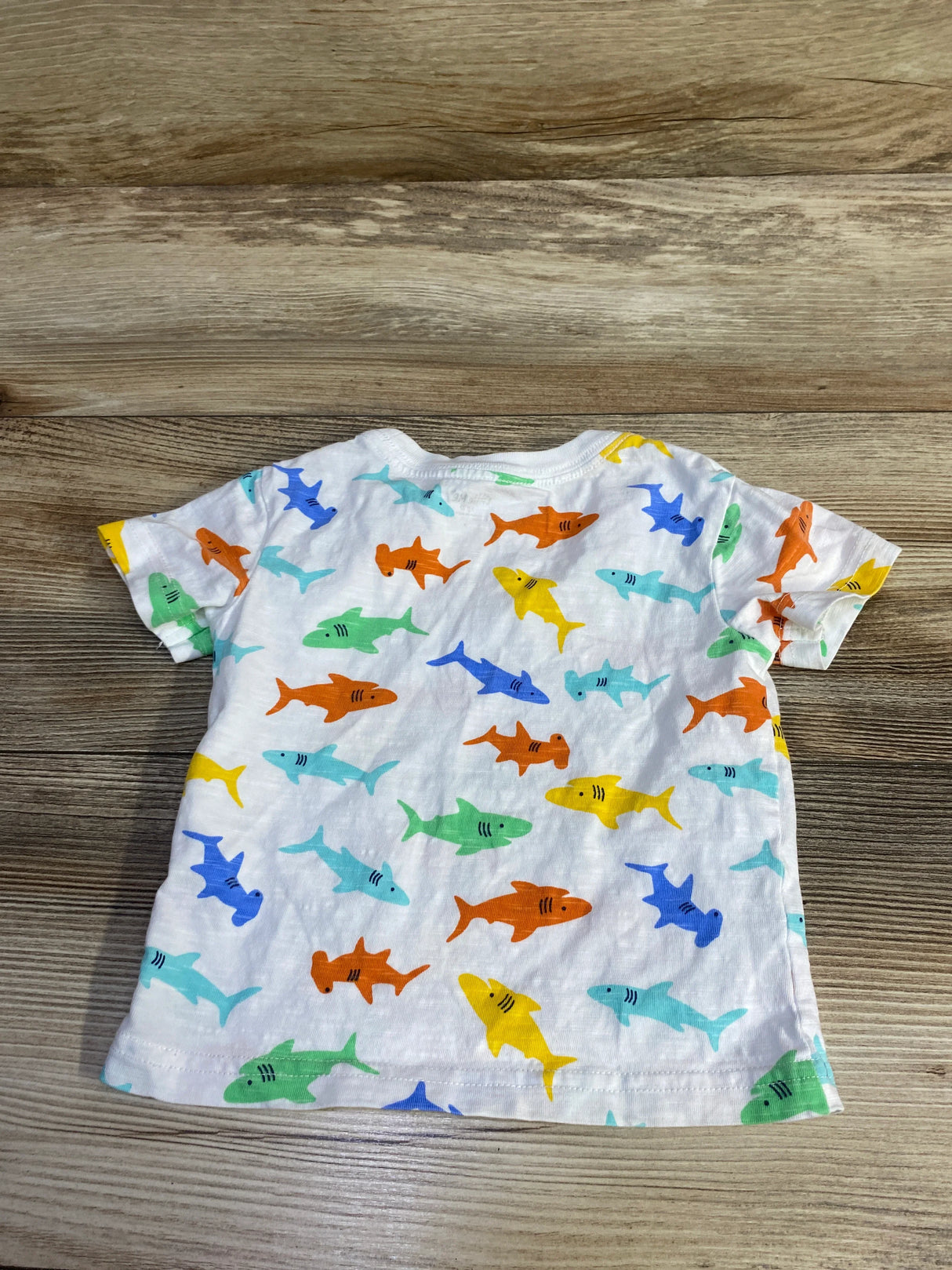 Flat lay of Little Me shark baby shirt size 12 months showing back view with multicolored shark print on white fabric over wood background