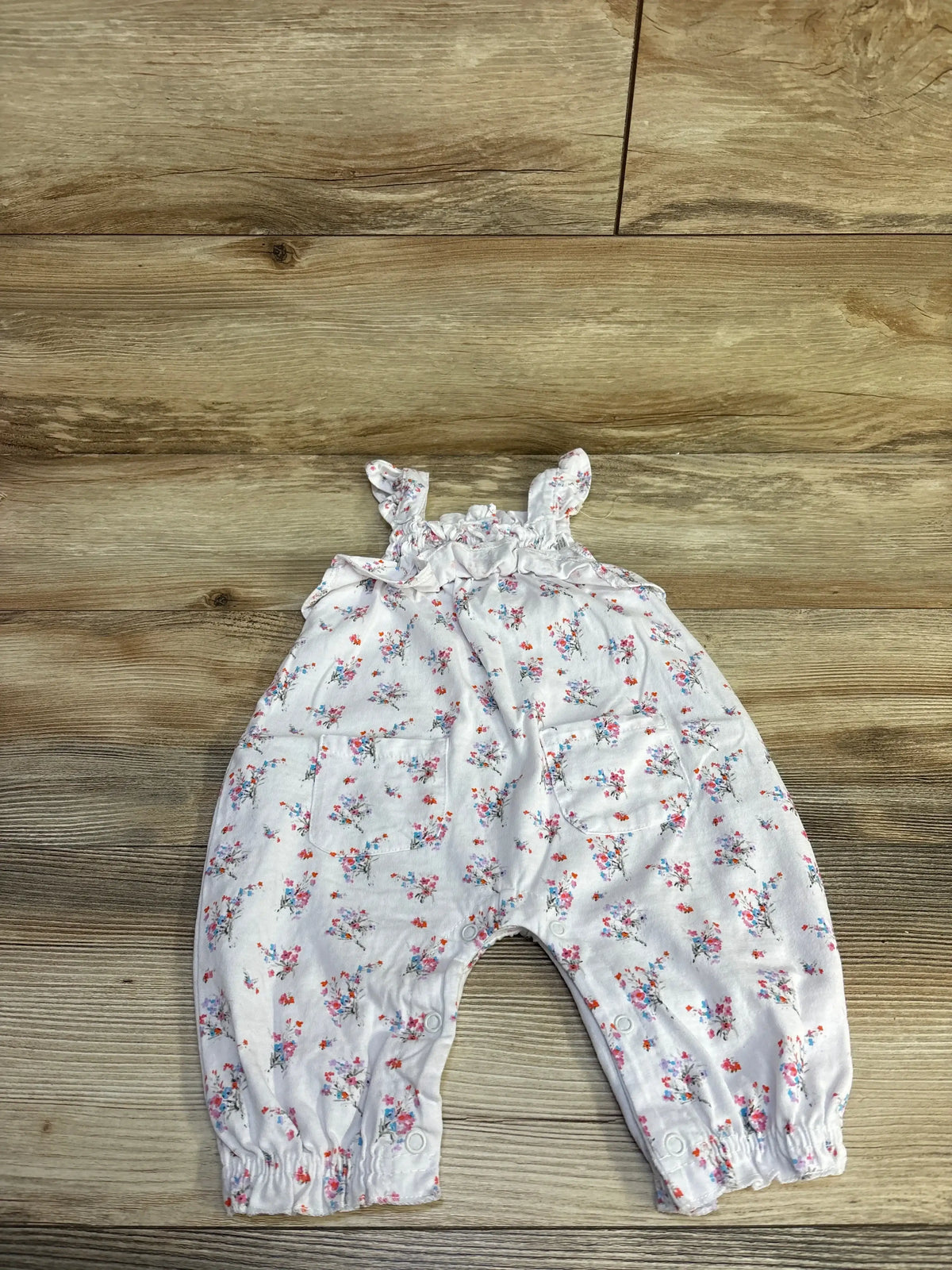 Top-down view of white Nannette baby jumpsuit 6-9M with floral print, ruffle straps, pockets and snap closures on wood floor