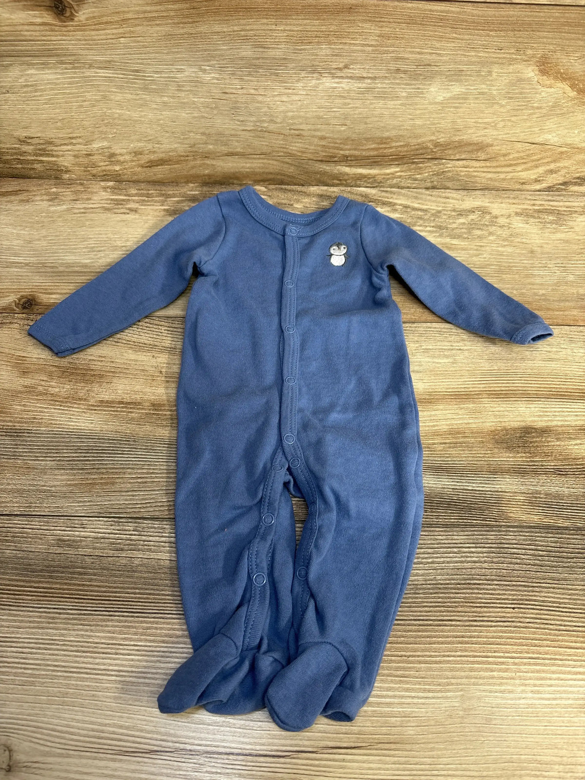 Flat lay of blue footed baby sleeper (3–6 months) showcasing front snap closure and penguin chest embroidery on wood background
