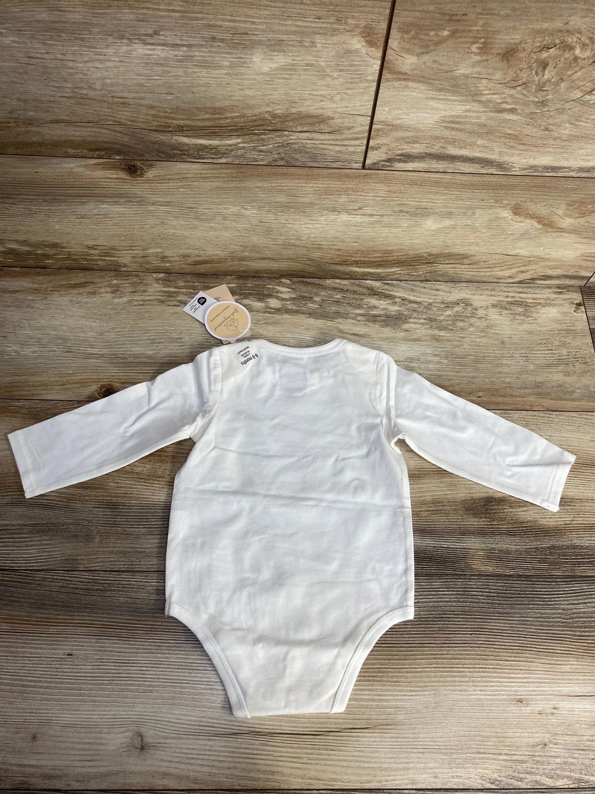 Plain white long-sleeve baby onesie laid flat on wooden floor with attached tags