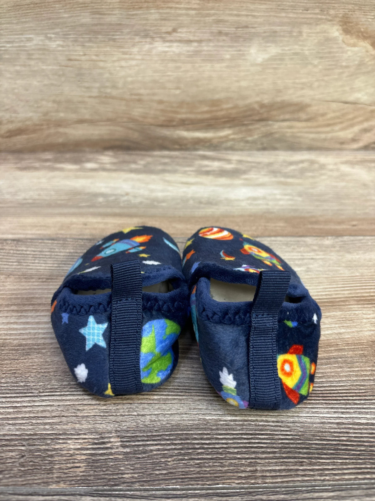 Rear view of Soft Cozy Space baby slippers showing navy space-print rockets, planets and stars with heel pull tabs on wooden surface