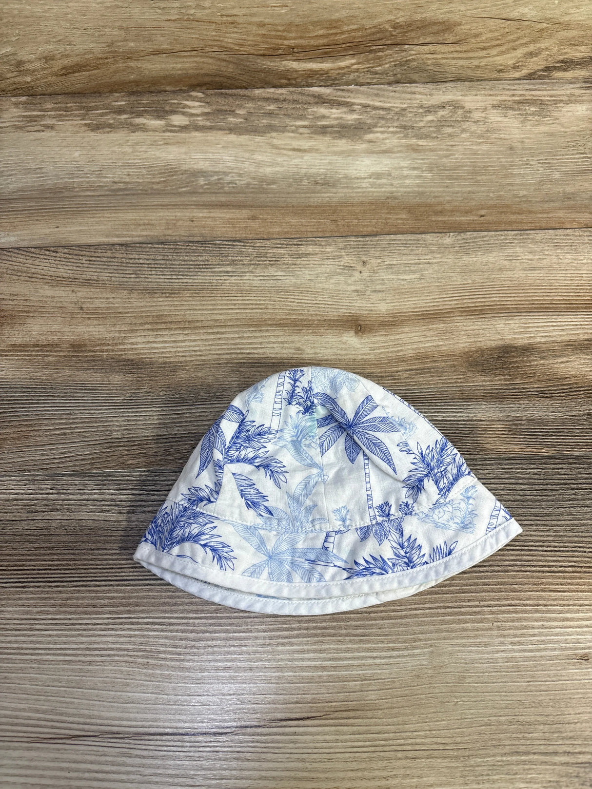 Top-down view of Janie & Jack bucket hat, size 0-3M, showing blue palm print on white fabric on wood background