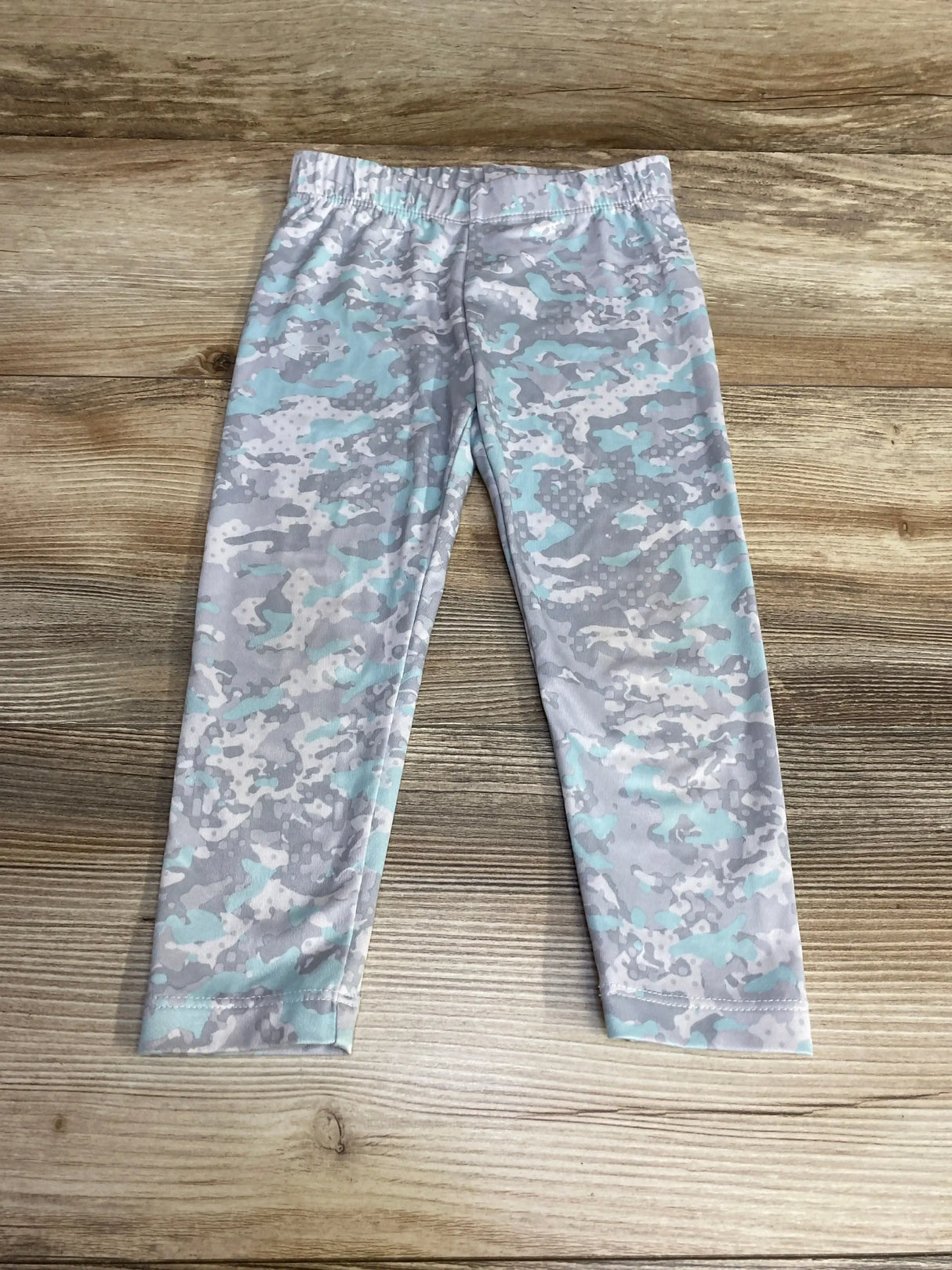 Flat lay of toddler Under Armour leggings in grey and mint camouflage showing elastic waistband and full-length legs on wood floor background