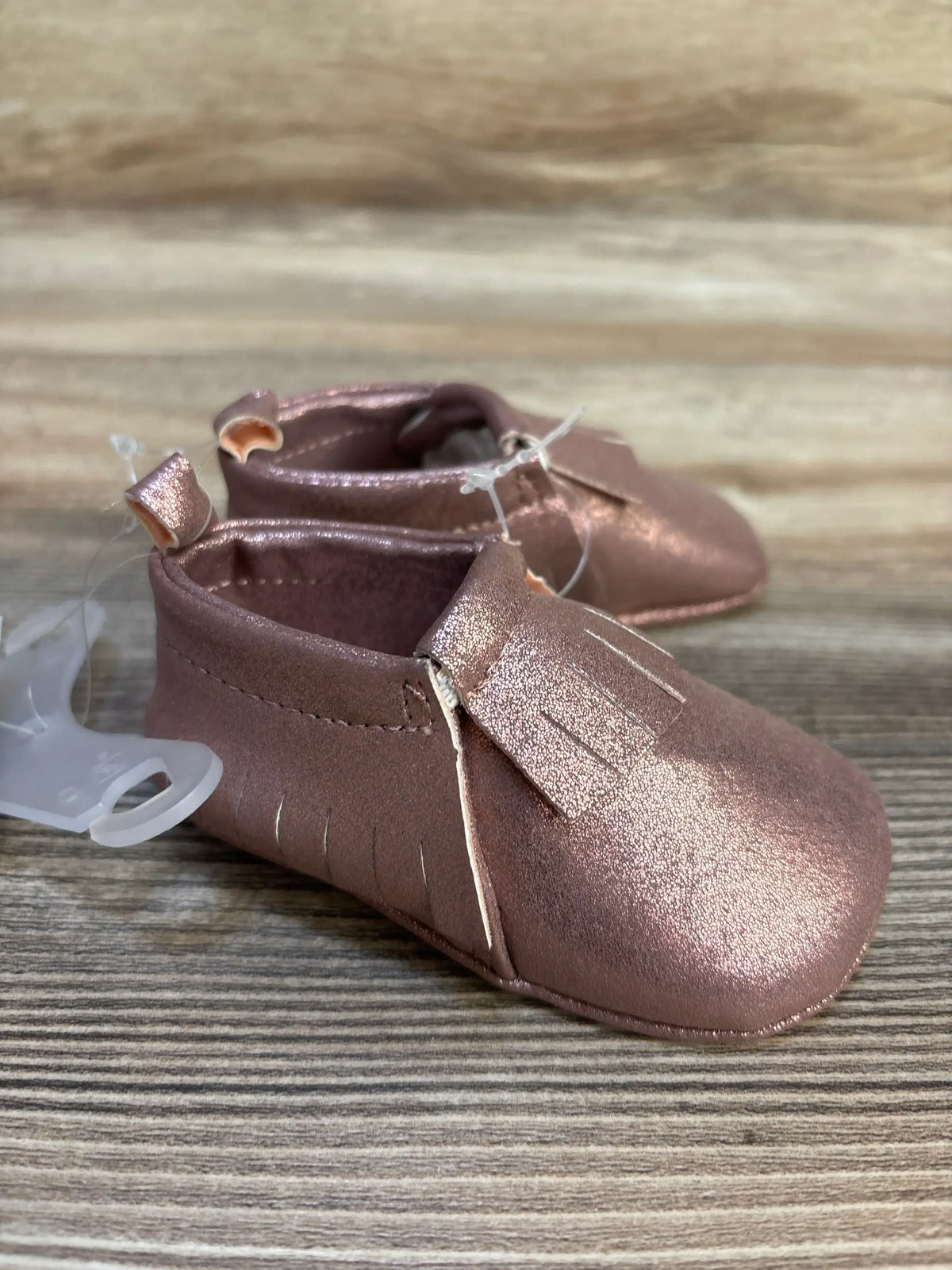 Metallic pink baby moccasins with fringe detail on wooden surface, infant footwear