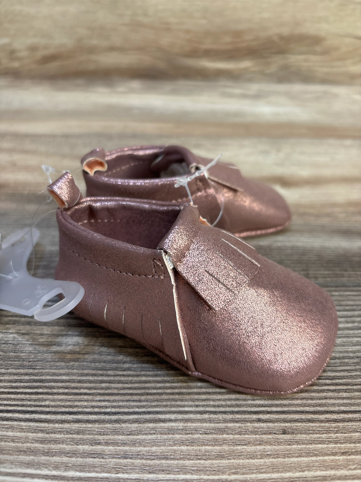 Metallic pink baby moccasins with fringe detail on wooden surface, infant footwear