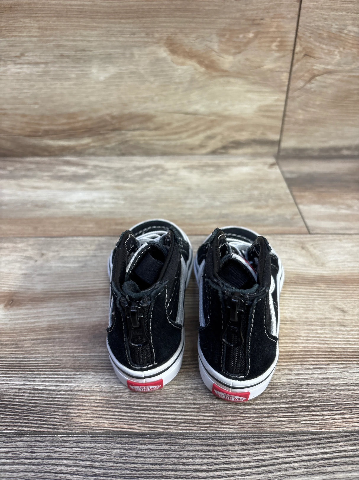 Rear view of black Vans Sk8-Hi zip toddler sneakers with back zipper, white side stripe and red heel tab on wood background
