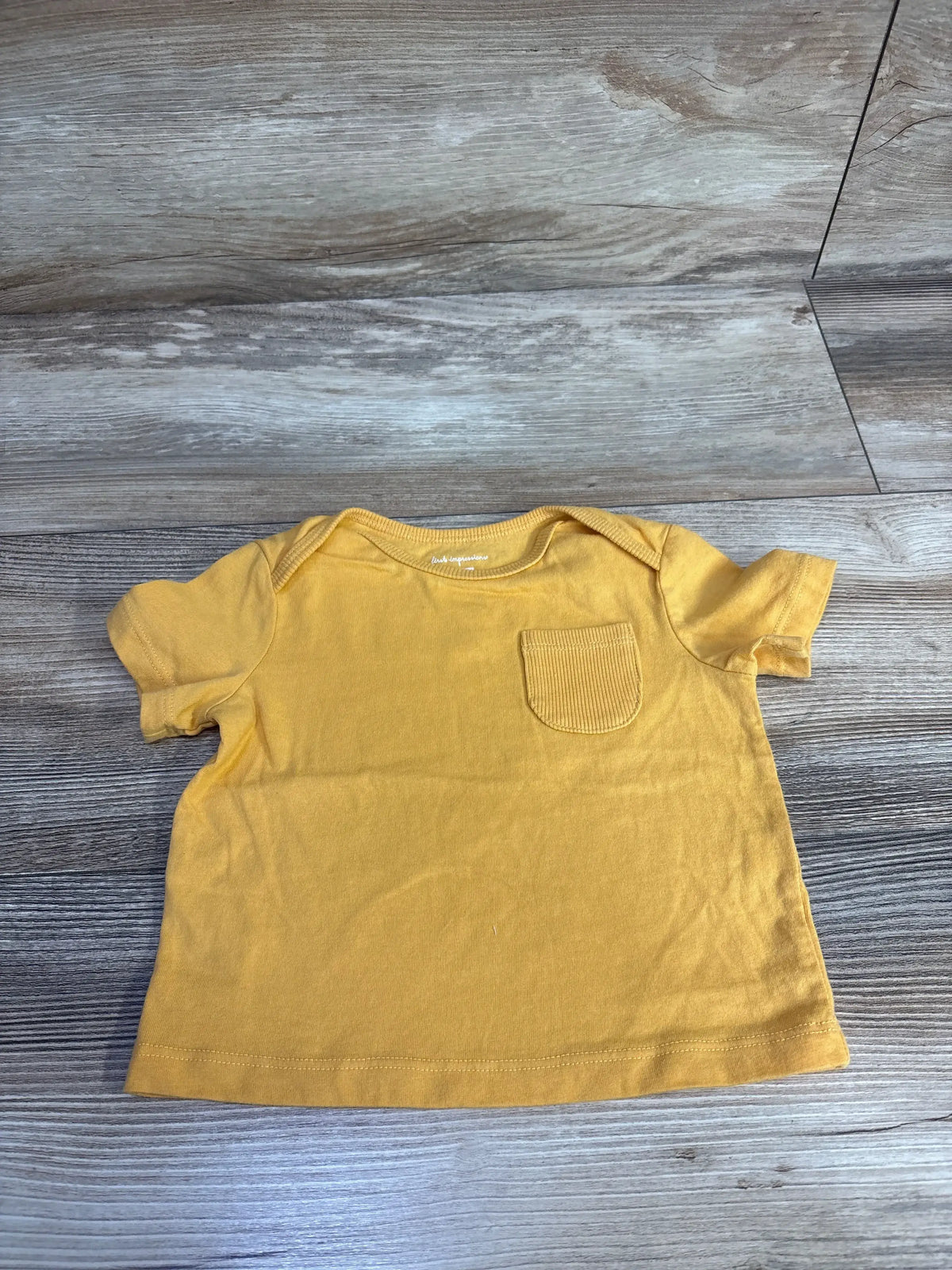 Flat lay of yellow baby pocket T-shirt size 6 to 9 months showing ribbed chest pocket and short sleeves on wood floor