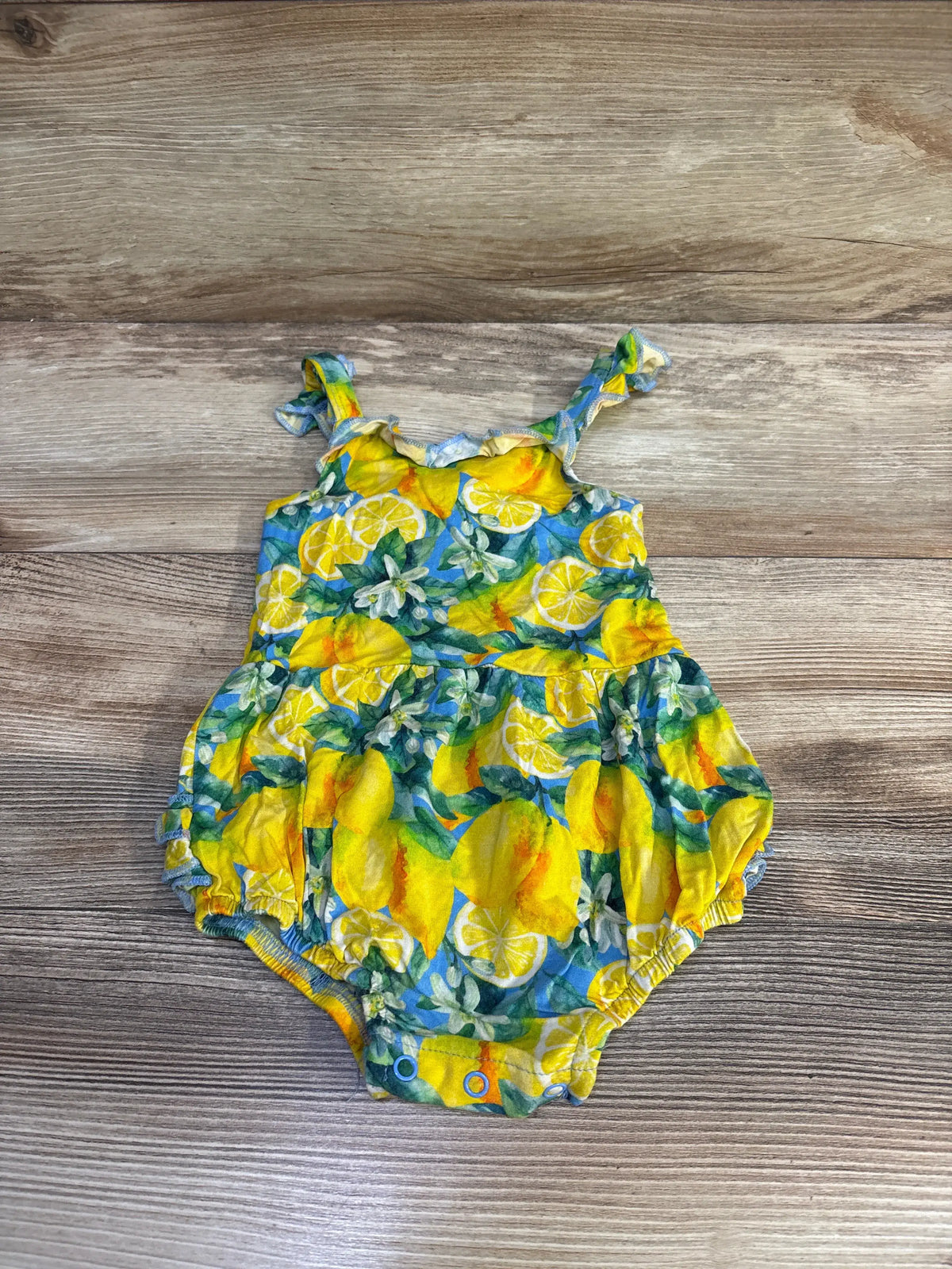 Flat lay view of Little Bum Bums lemon print bamboo shortie romper showcasing yellow lemon pattern and snap closures on a wood-look floor