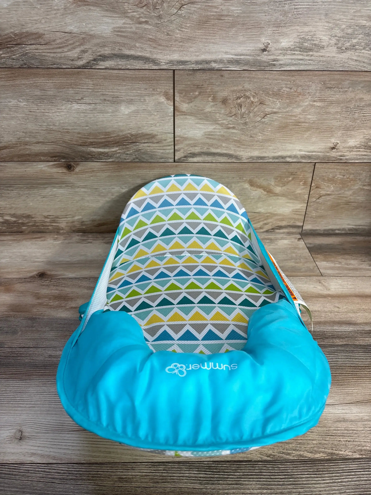 Front view of Summer Deluxe baby bather in teal with green and gray triangle print padded seat on wood plank floor