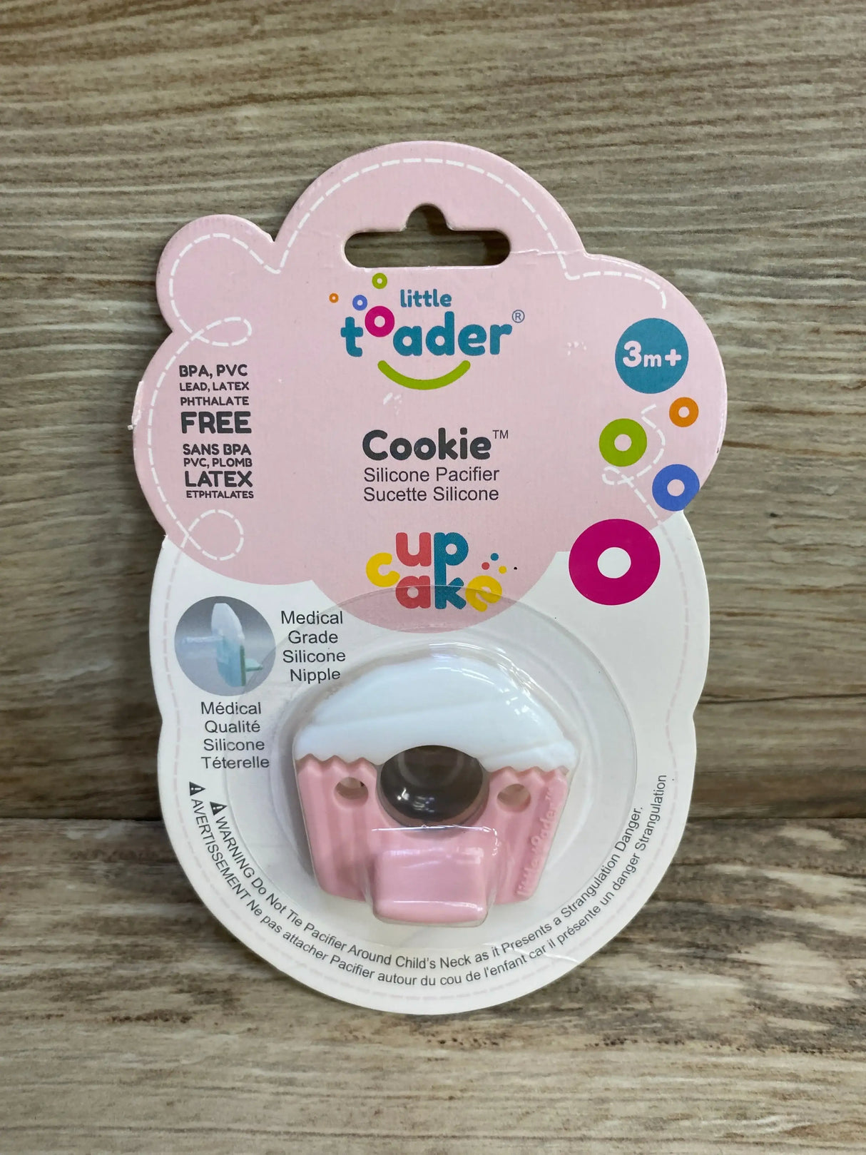 Front view of Little Toader Cookie cupcake pacifier showcasing pink silicone nipple and white top with packaging on wood background