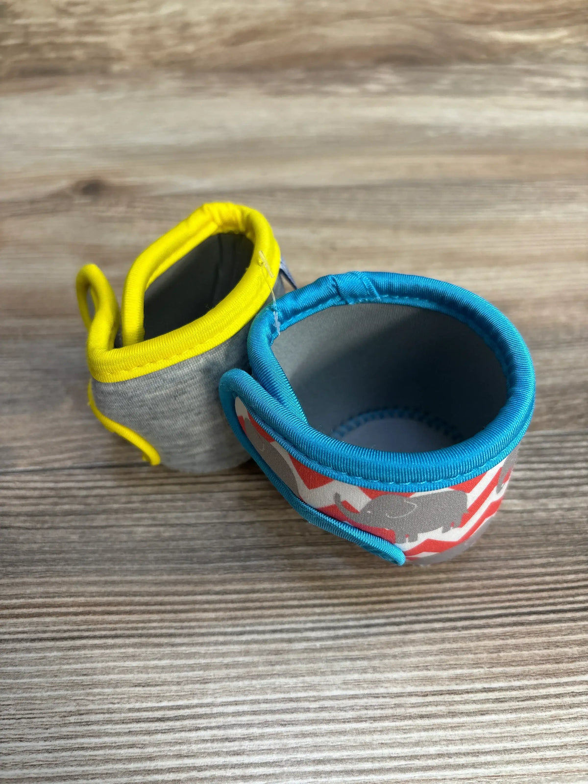 Close-up of Miracle Bean Neoprene Baby Bottle Sleeves 2pc one grey with yellow trim and one elephant print with teal trim on wood plank floor