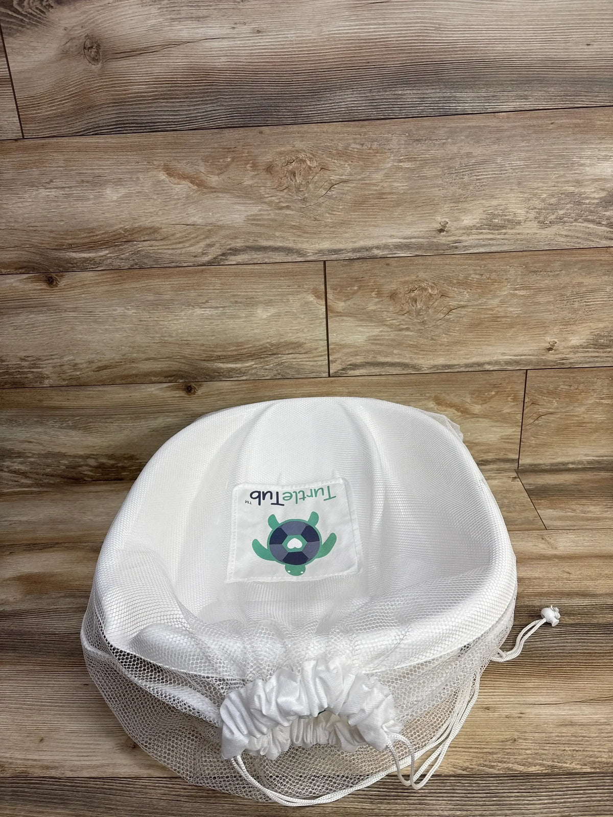 Top-down view of TurtleTub Swaddle Bath Tub showing white mesh, turtle logo patch and drawstring on wooden floor