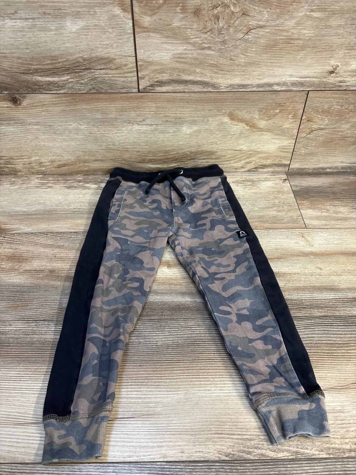 Kids camo jogger pants with black side stripes and drawstring, laid flat on wood floor