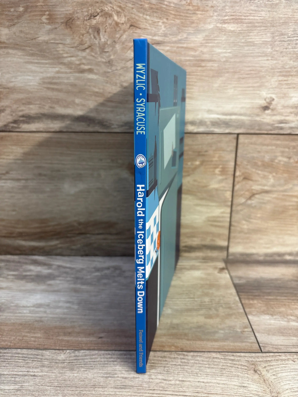 Side view of Harold the Iceberg Melts Down hardcover book showcasing its blue spine with a wood plank background
