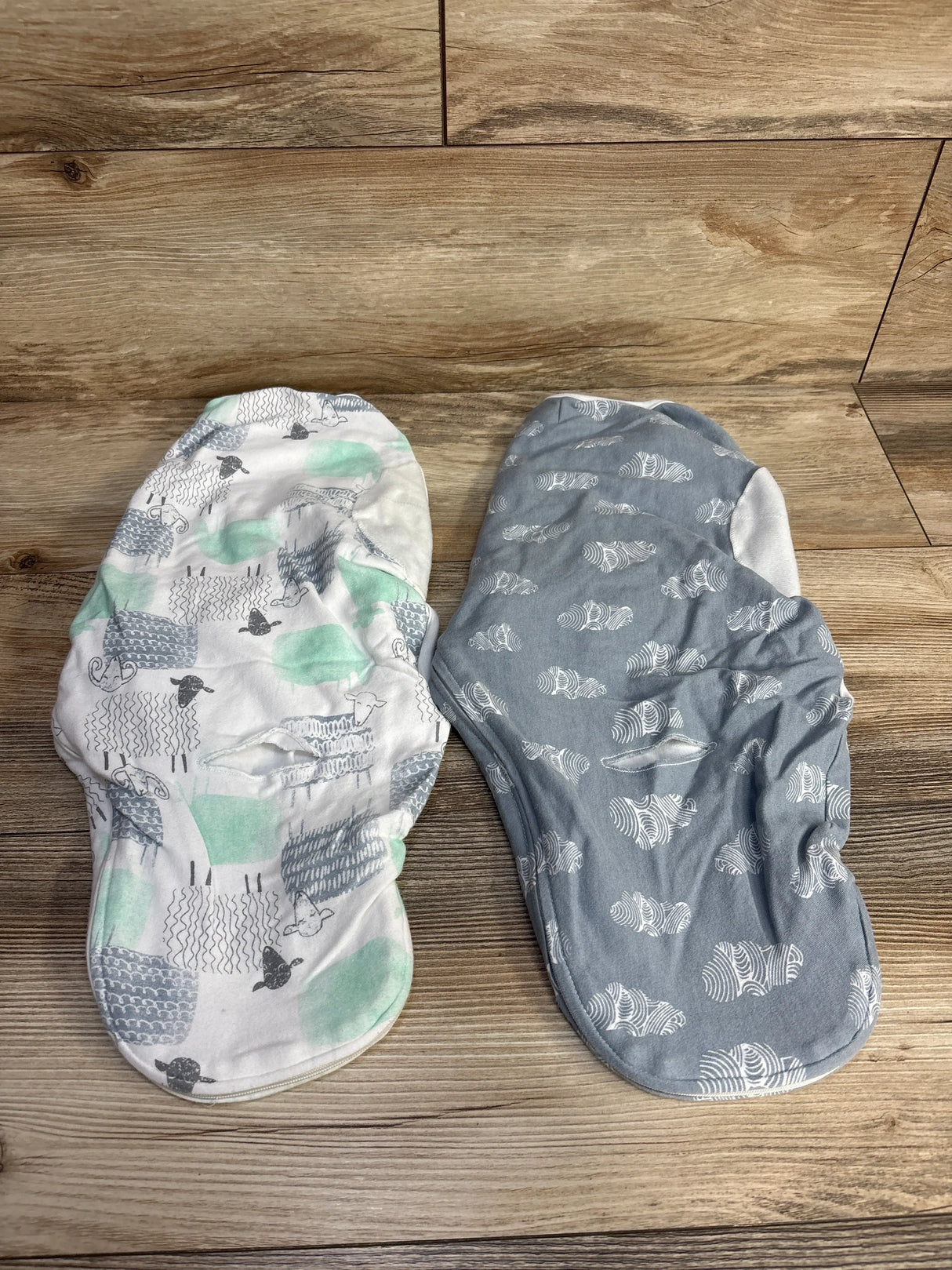 Top down view of two Aden + Anais newborn swaddle wraps, white sheep-print and gray cloud-print, contoured shape on wood background