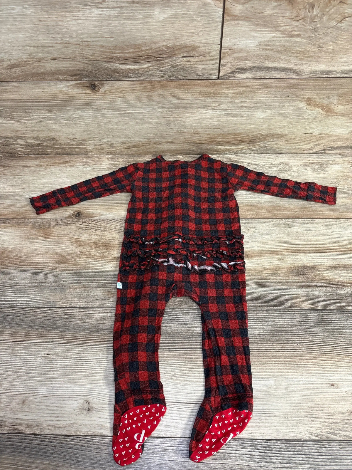 Red and black plaid baby sleeper with ruffle back and footed grips, laid on wood floor