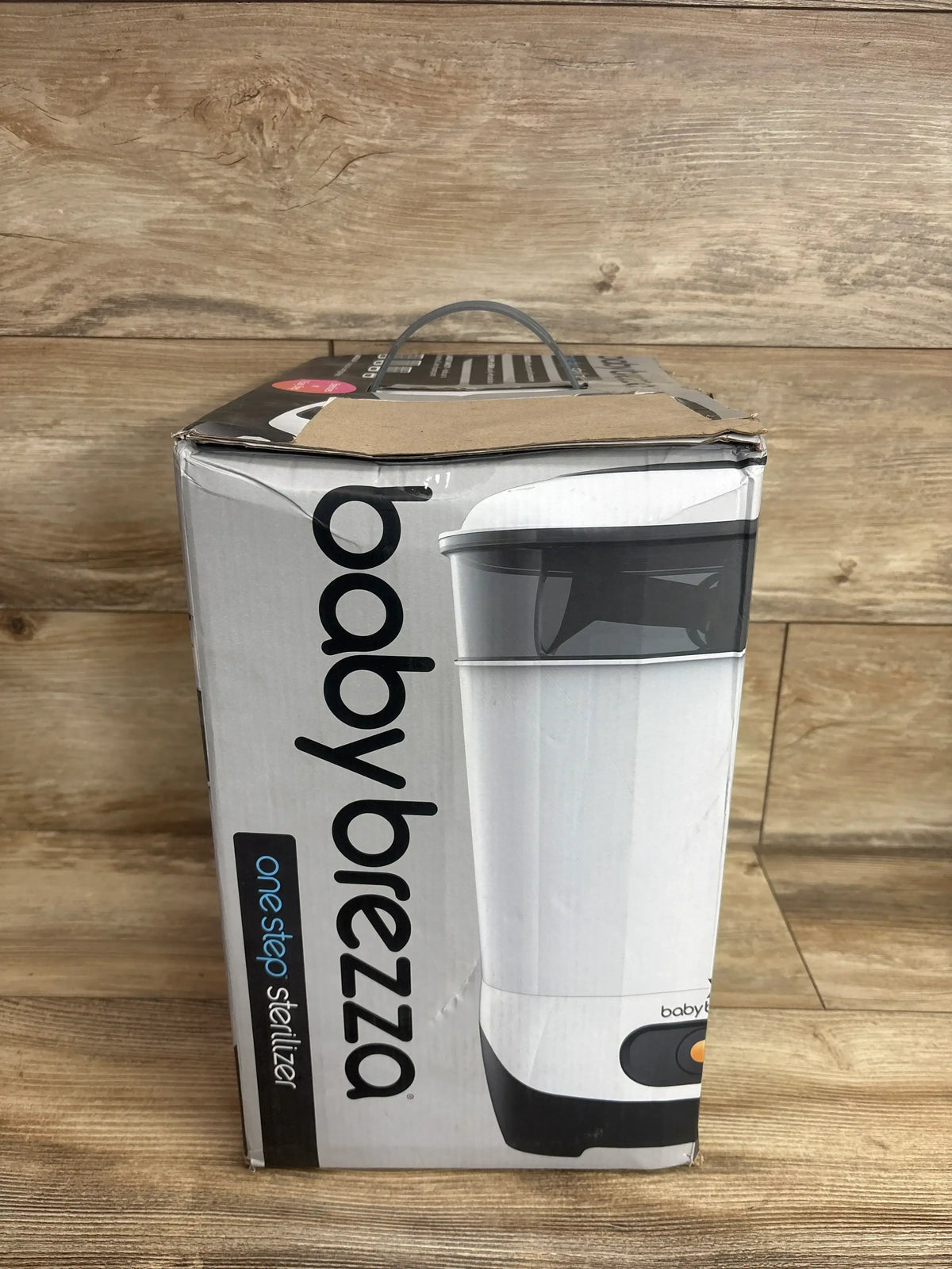 Front view of Baby Brezza One Step Sterilizer box showing product image and dented top handle on wood floor background