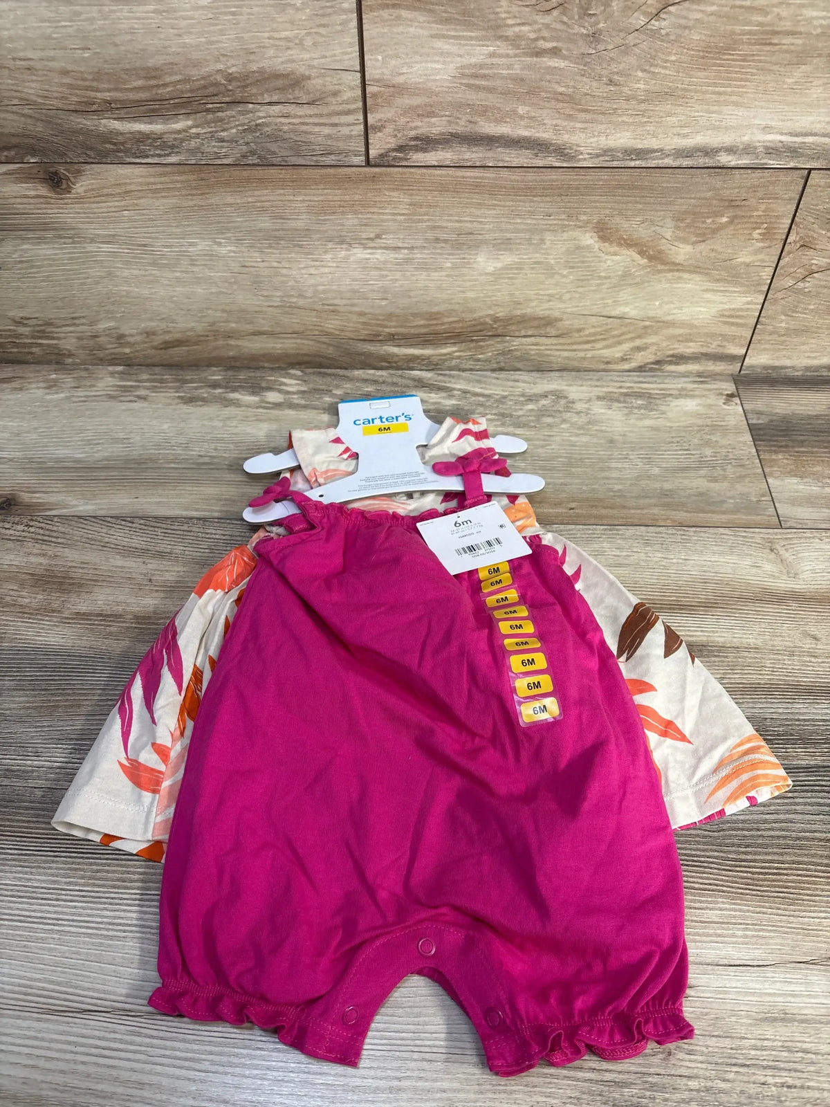 Top-down of Carter’s 3-piece 6M dress set showcasing bright pink romper with snap crotch and beige floral dress on wood floor