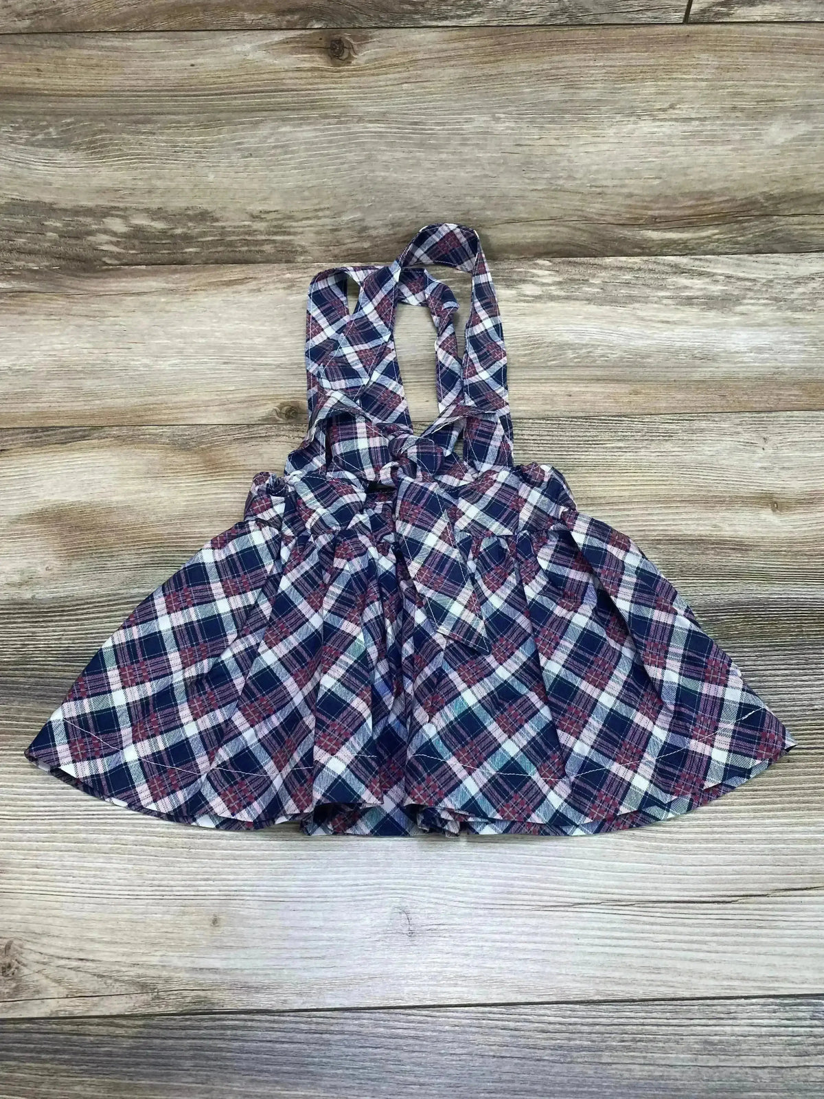 Flat-lay of baby suspender plaid skirt showcasing navy and pink plaid, crossed shoulder straps, gathered waist, and flared hem on wood background
