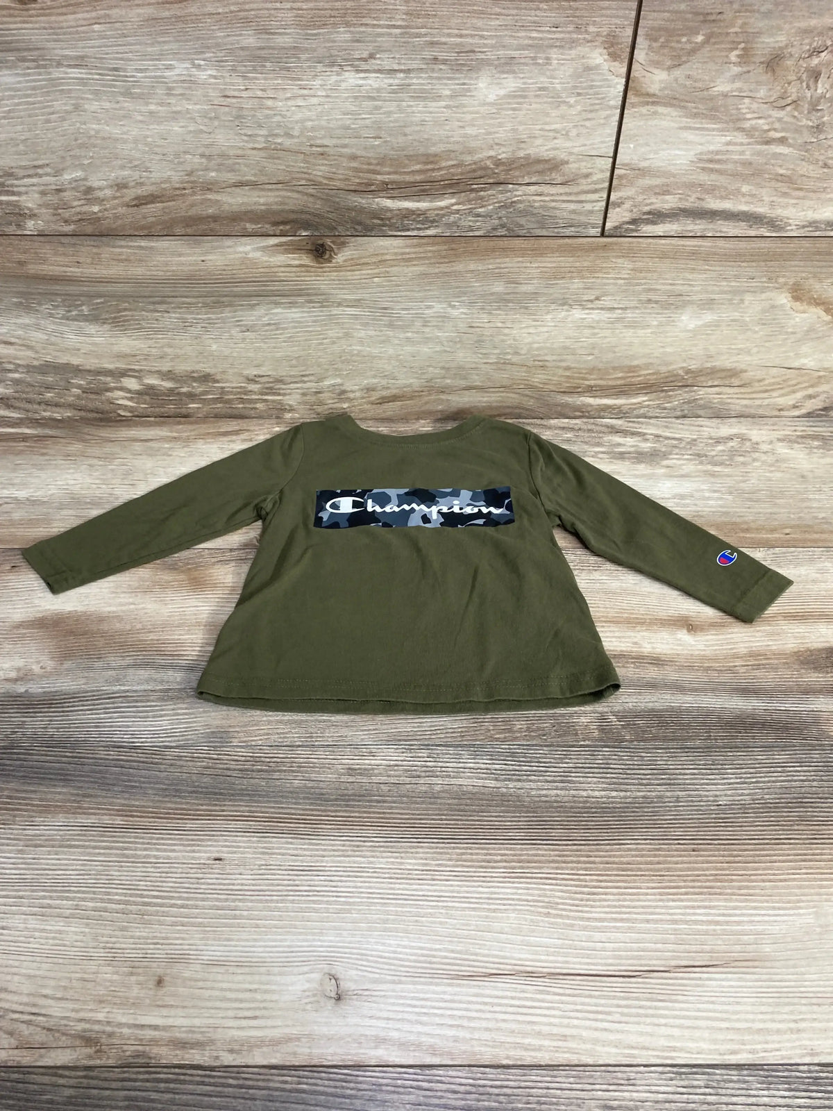 Front view of green Champion long-sleeve shirt showcasing camouflage logo and C sleeve patch on wood background