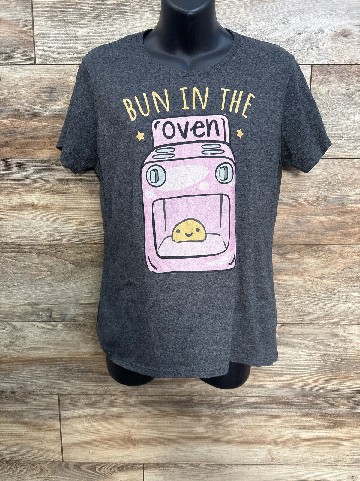 Front view of gray maternity shirt with short sleeves, pink oven graphic, and Bun in the Oven text on wood background