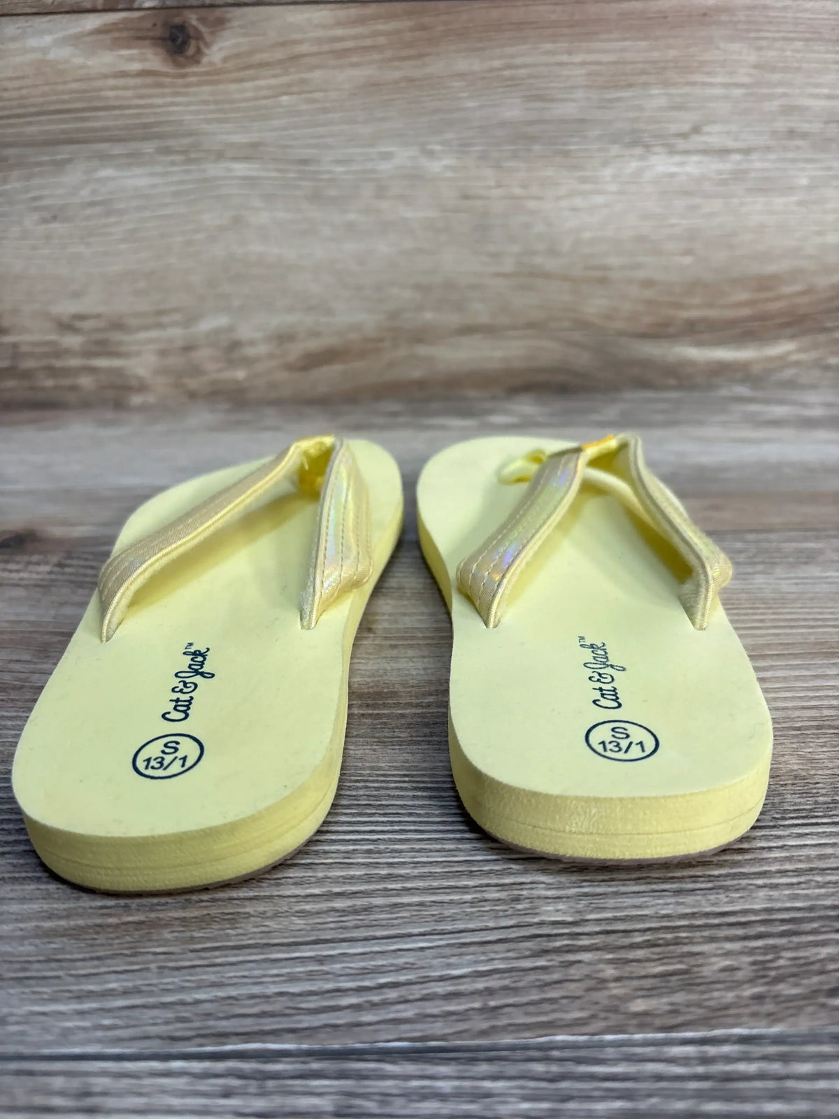 Front view of Cat & Jack Ava kids’ yellow flip flops showcasing glitter straps and printed size S 13/1 on wood background