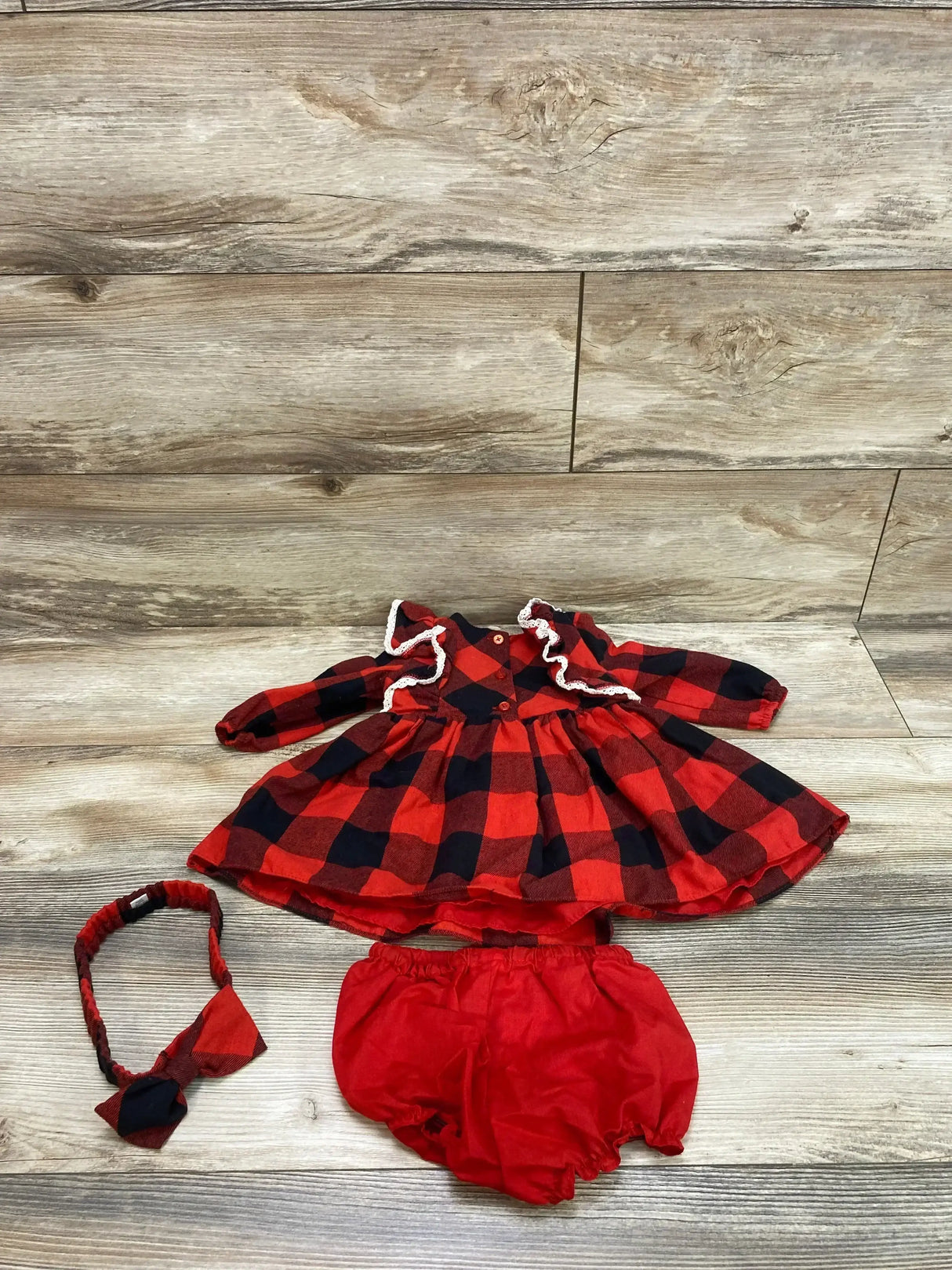 Flat lay of 3-piece red and black buffalo check baby dress set with ruffled lace collar, matching headband and red bloomers on wood backdrop