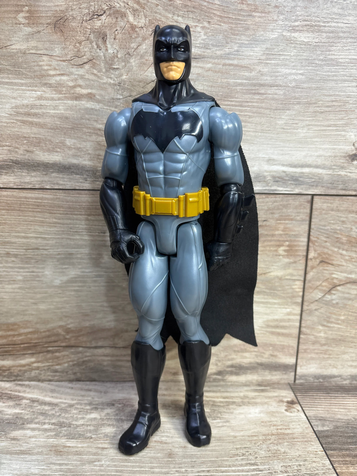 Batman action figure toy with gray suit, black cape, and yellow utility belt standing on wooden surface