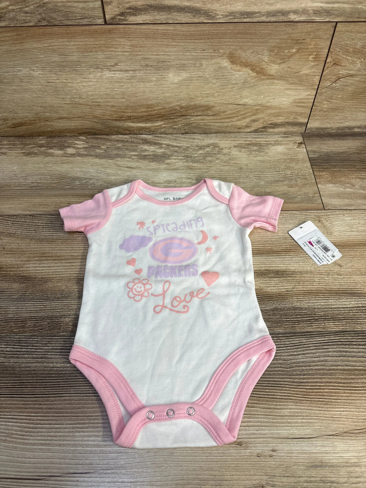 Front view of NFL baby bodysuit showcasing Packers logo and Spreading Love design with pink trim, laid flat on wood-grain flooring