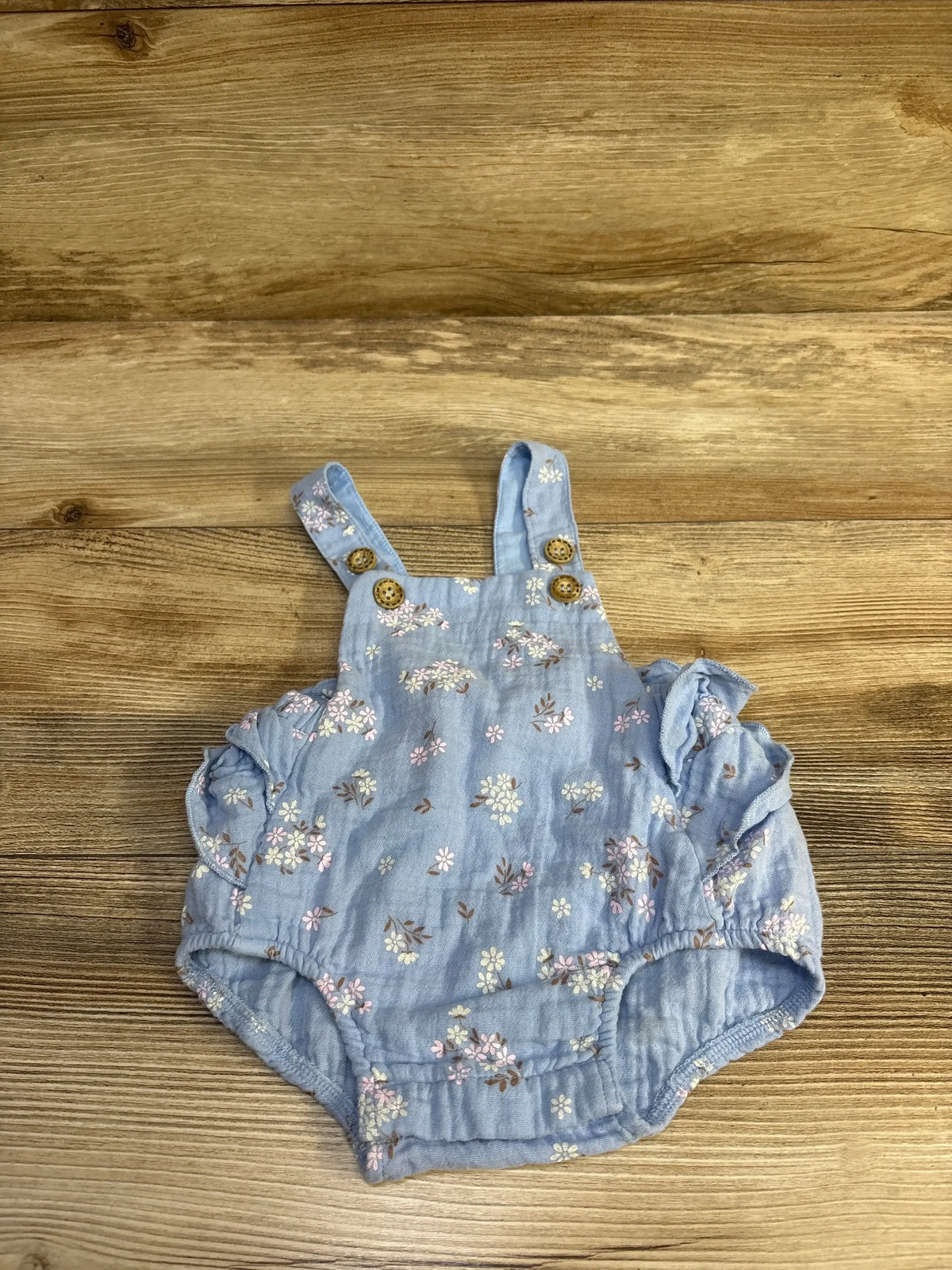 Flat lay of Purebaby Organic floral ruffle bubble romper size 0-3m in light blue with floral print wooden buttons and ruffle leg openings on wood plank floor