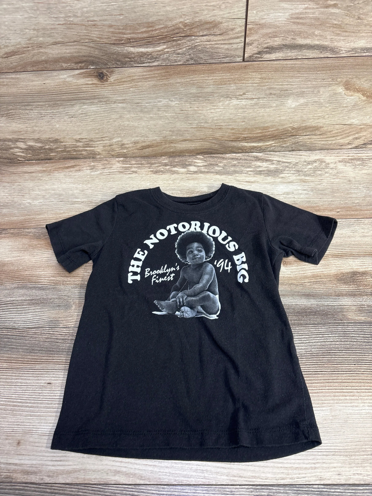 Black toddler T-shirt with white graphic of baby and text The Notorious B.I.G. Brooklyn's Finest '94 on wood floor