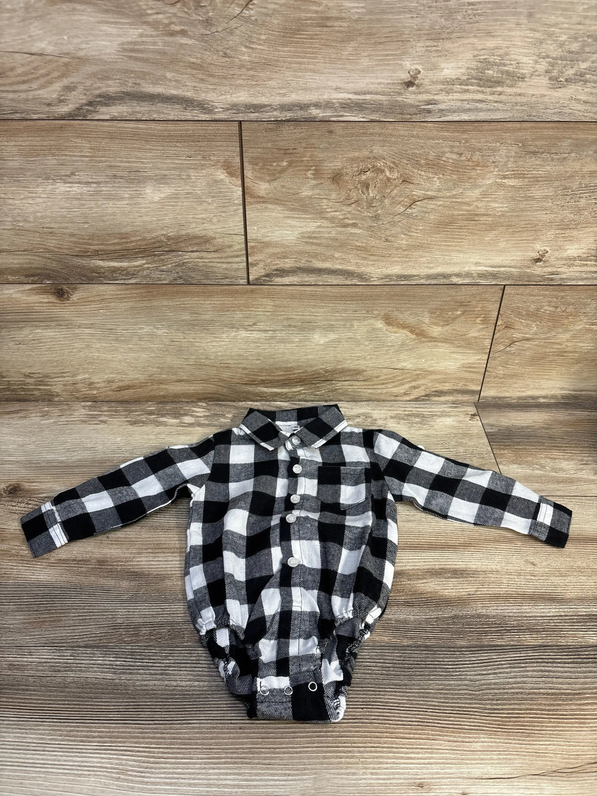 Front view of a black plaid flannel bodysuit showcasing long sleeves, collar, buttons, and snap closures on a wood-look background