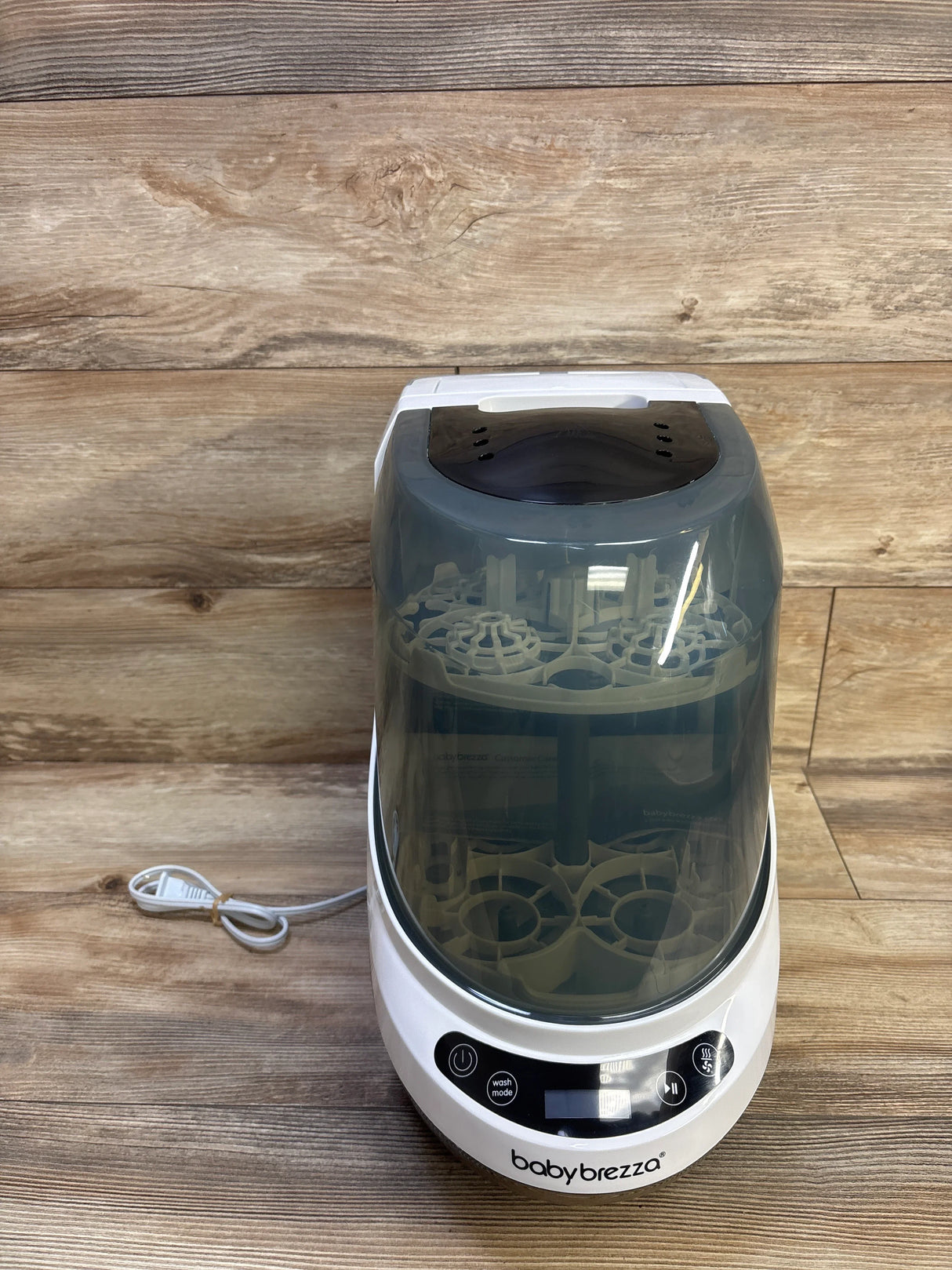 Front view of Baby Brezza Bottle Washer Pro showing translucent lid revealing bottle racks and front control panel on wood background