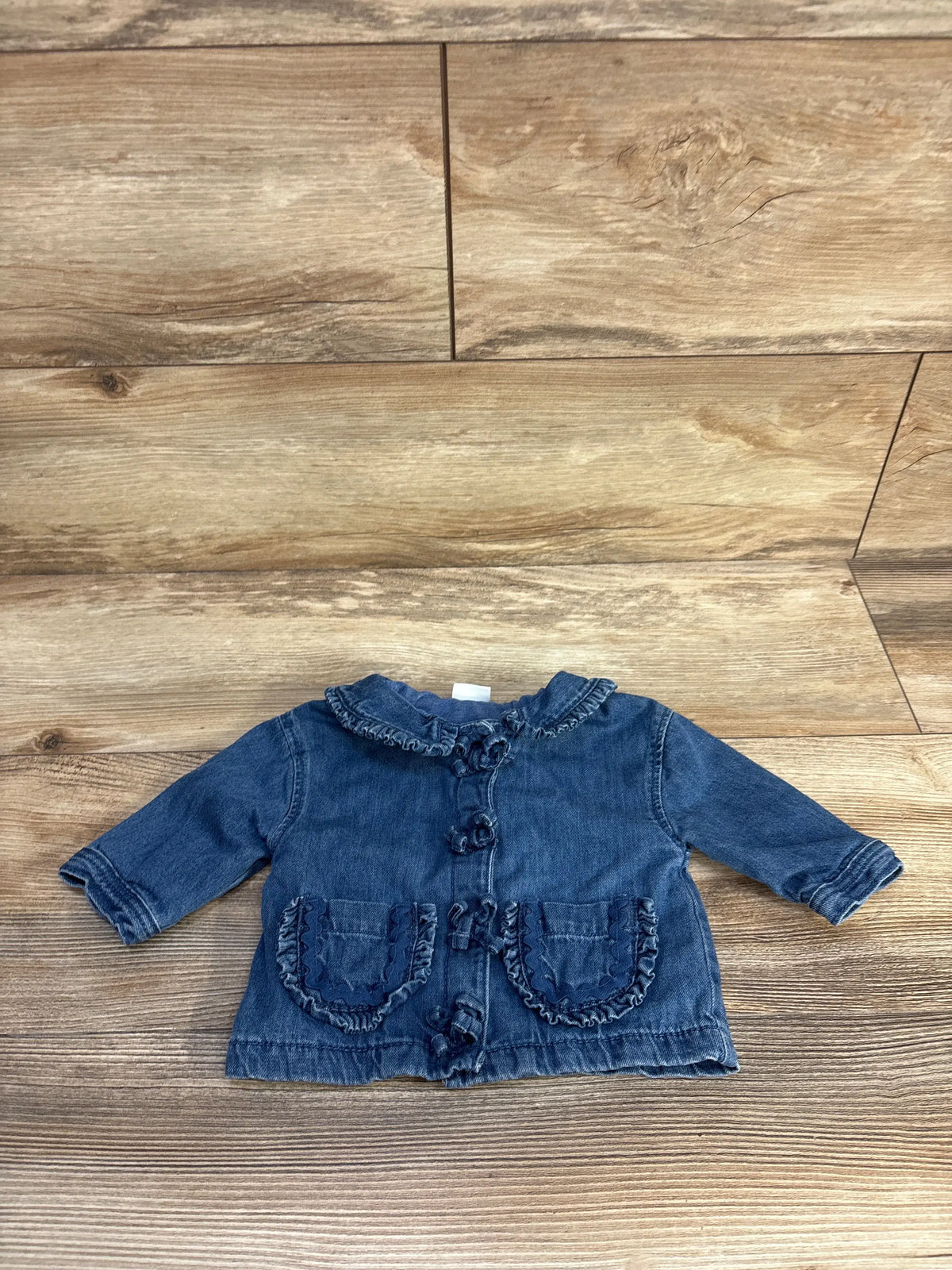Front view of Next blue denim jacket showcasing ruffled collar, button front, and two pockets on wood-look flooring