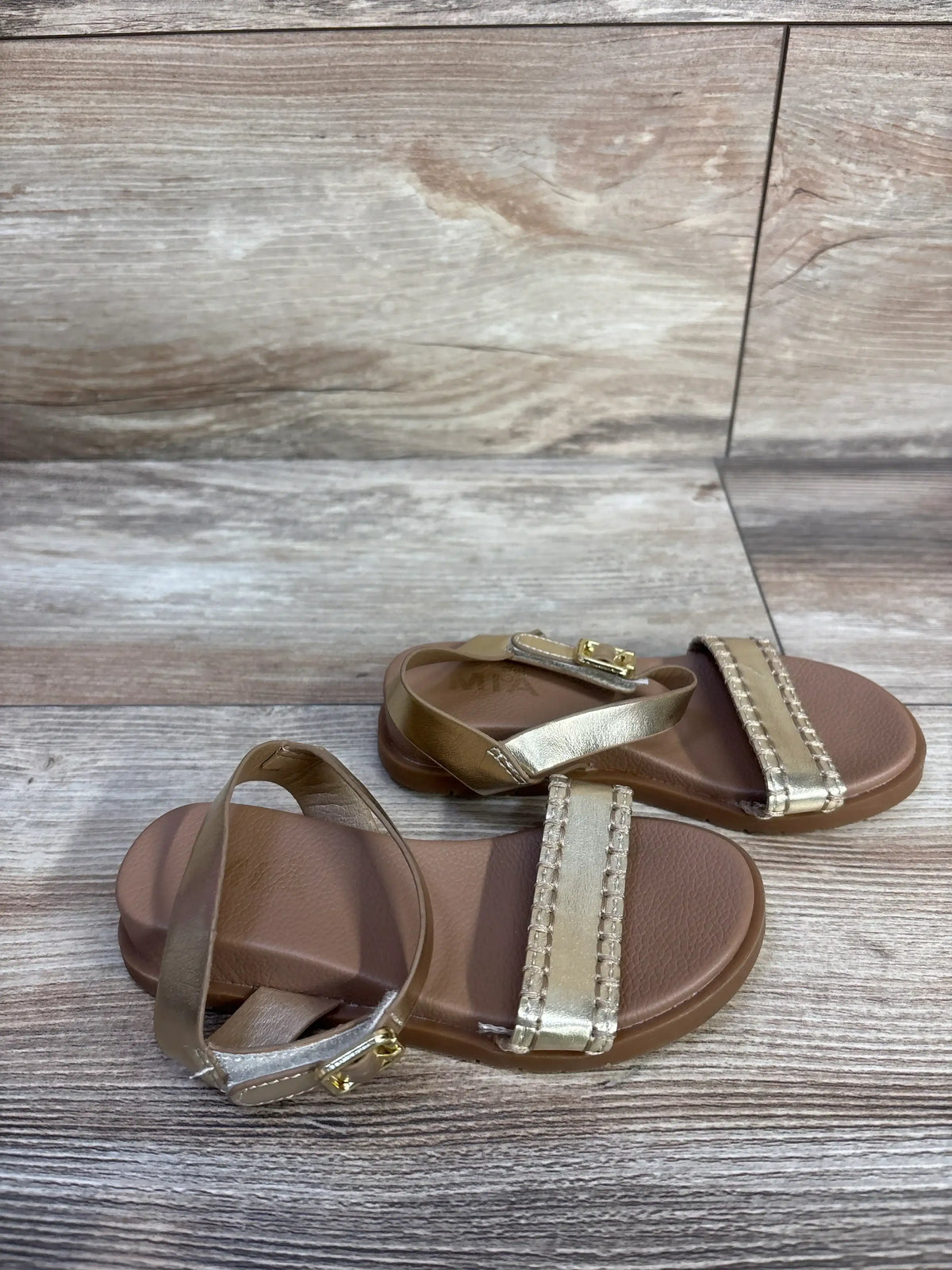 Angled top view of a pair of MIA Peyton sandals showing gold metallic straps and braided toe band on light wood background